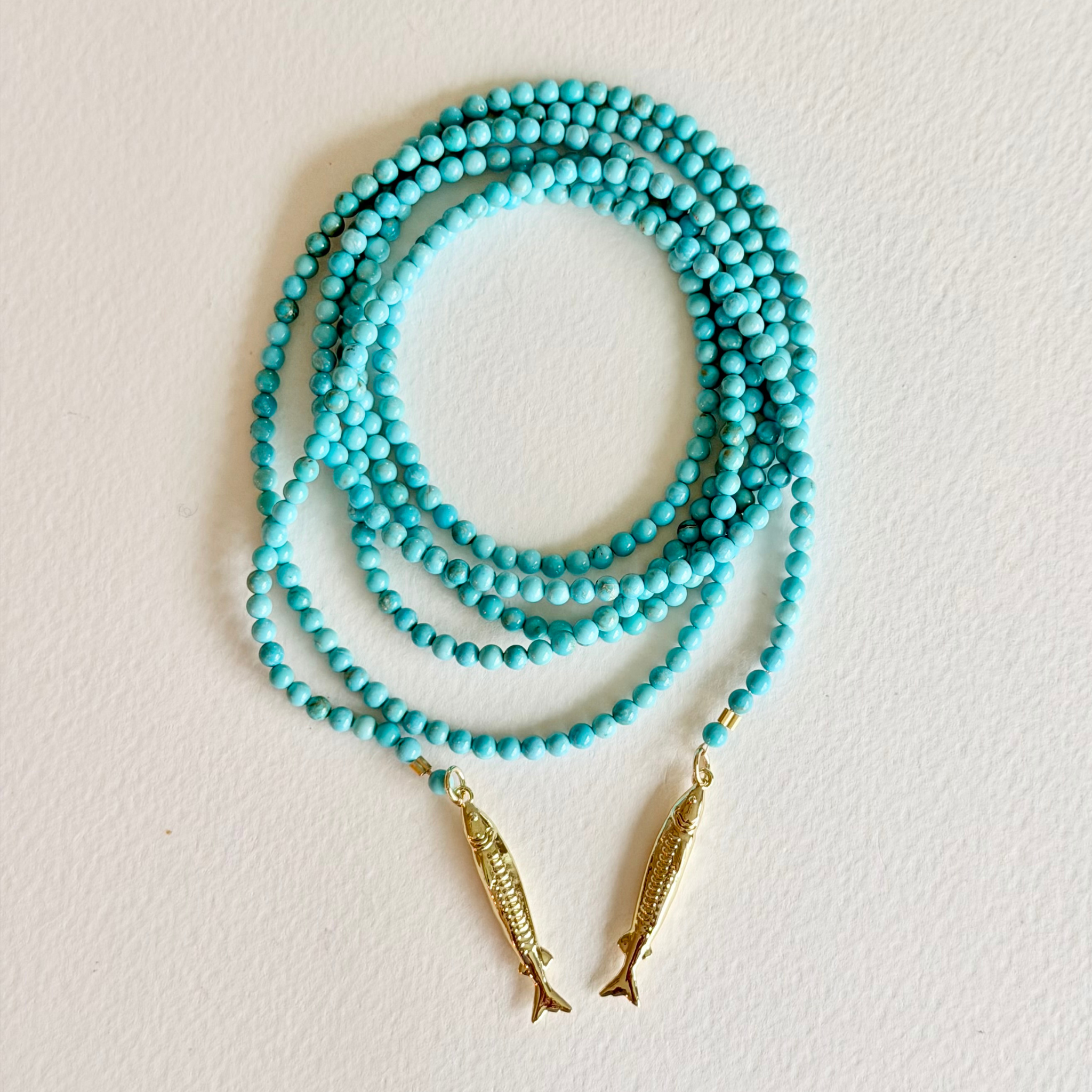 Turquoise beaded necklace with gold fish pendants on a light beige background