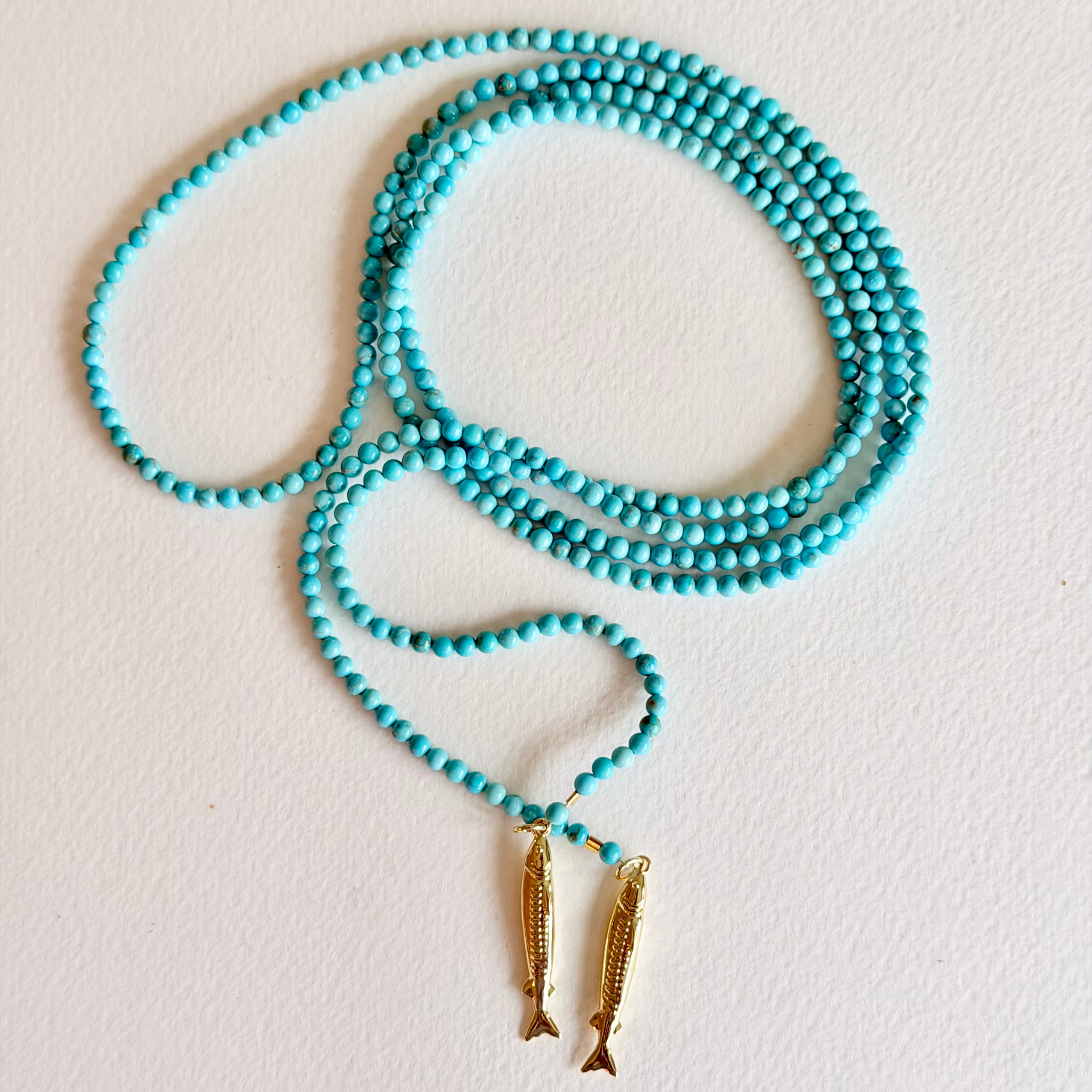 Turquoise beaded necklace with gold fish pendants on a light gray background