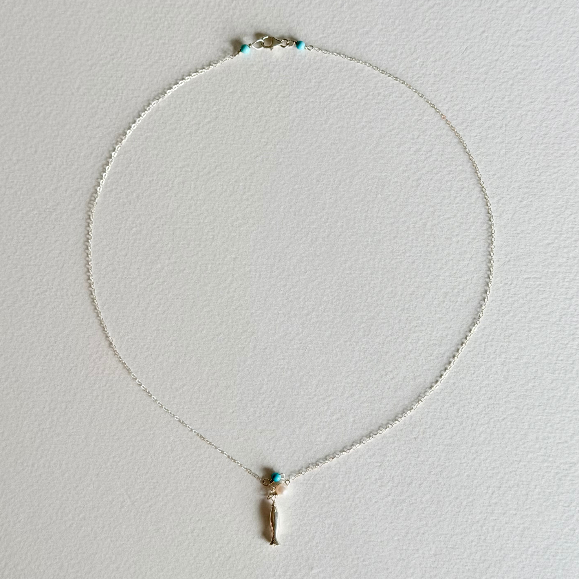 A delicate silver chain necklace with a small gold-toned pendant featuring a tiny turquoise blue bead accent, displayed on a textured off-white background.