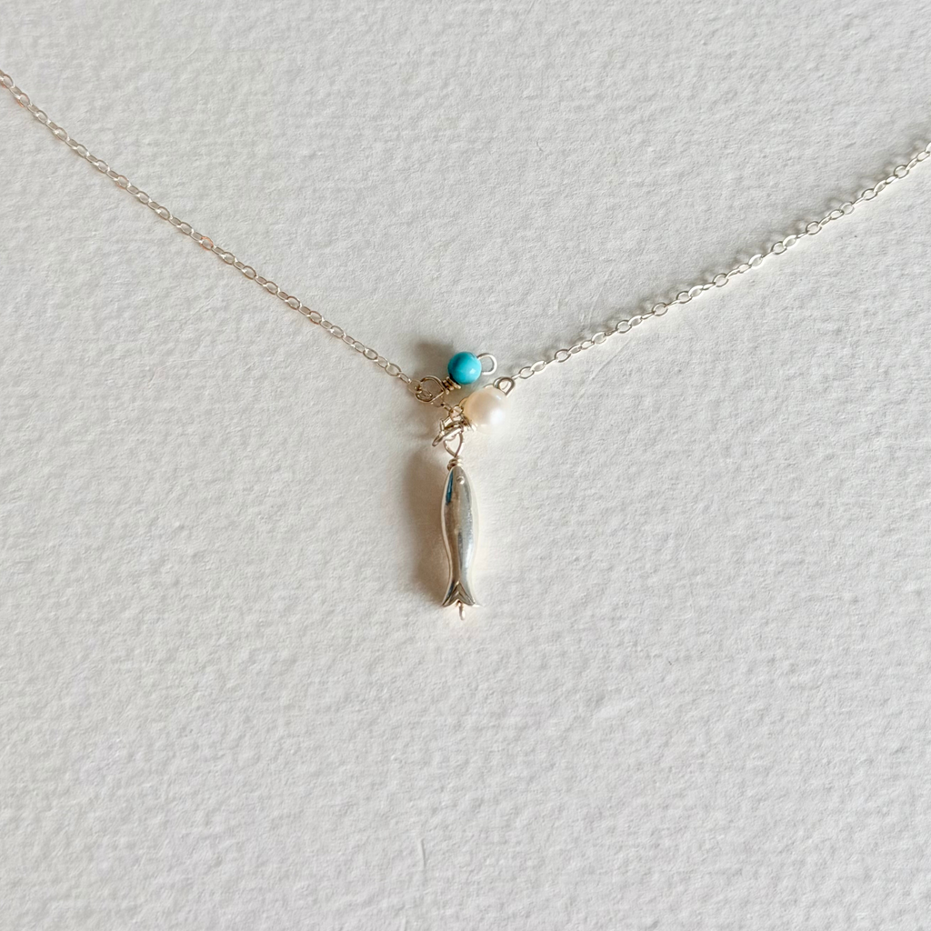 A delicate silver chain necklace featuring a small fish charm wrapped in silver wire, accented by a tiny turquoise bead and a white pearl, displayed on a textured white background.