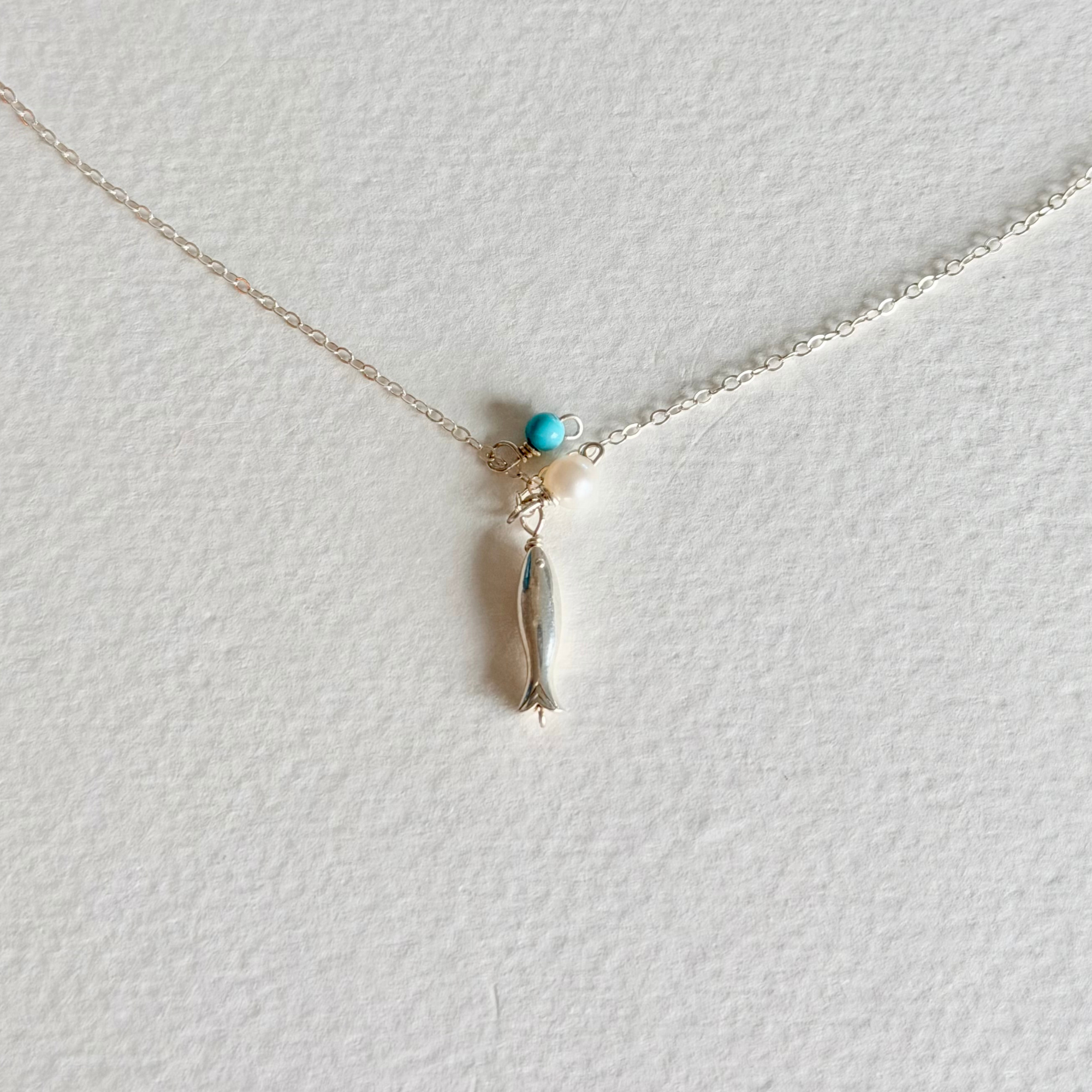 A delicate silver chain necklace featuring a small fish charm wrapped in silver wire, accented by a tiny turquoise bead and a white pearl, displayed on a textured white background.