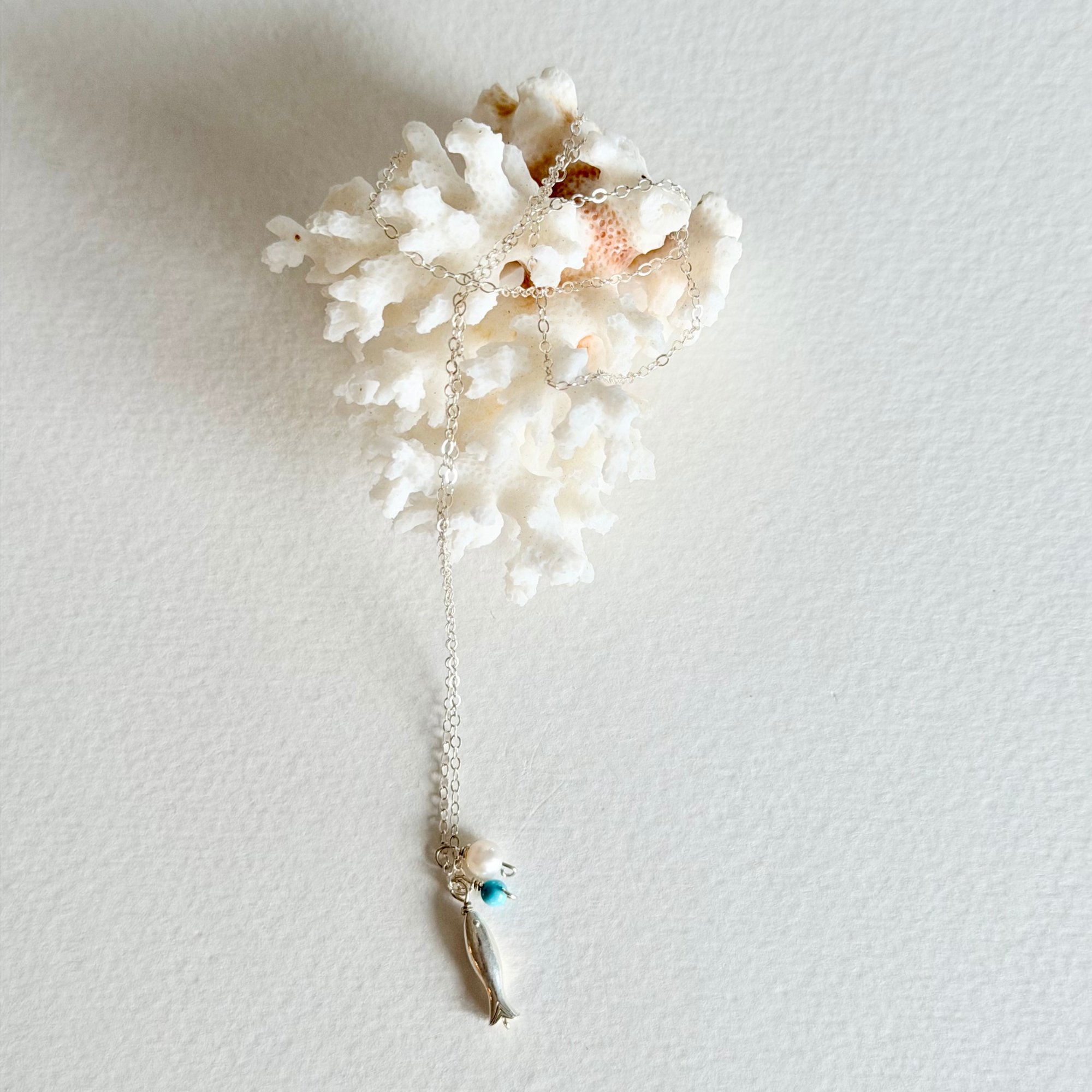 A seashell and coral-inspired pendant necklace with a silver chain and a small turquoise bead charm, displayed against a textured off-white background.