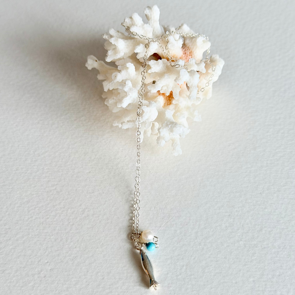 A silver-toned necklace with a small fish charm and a turquoise bead, displayed draped over a white coral shell on a textured off-white background.