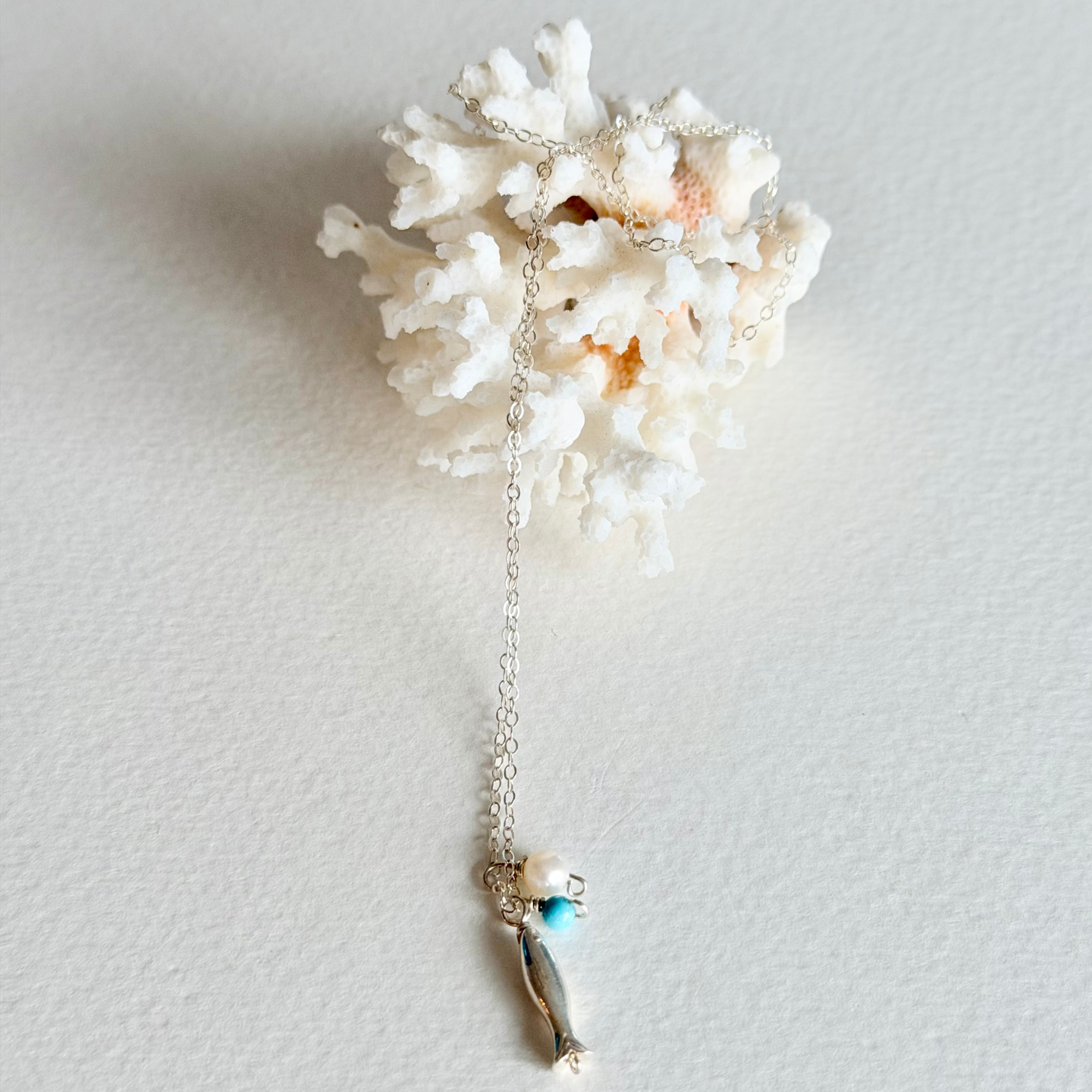 A silver-toned necklace with a small fish charm and a turquoise bead, displayed draped over a white coral shell on a textured off-white background.