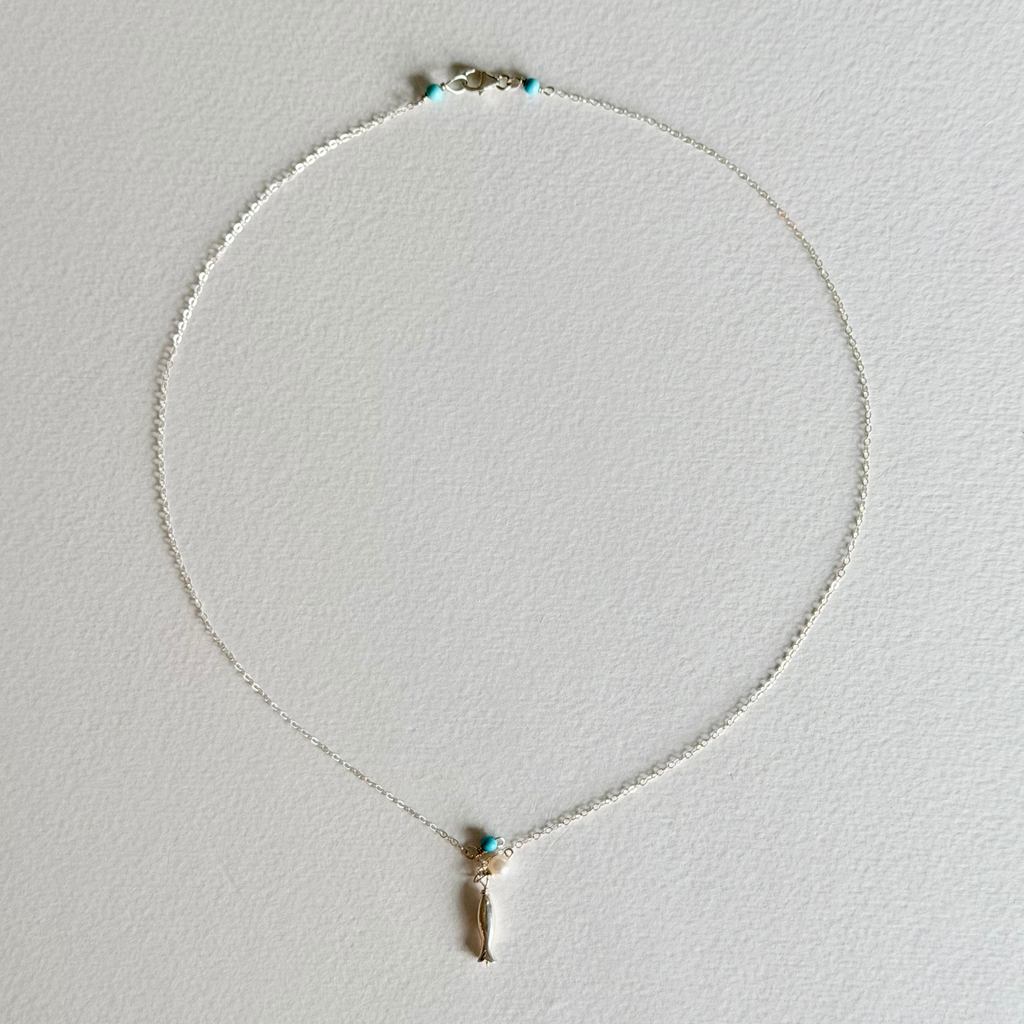 A delicate silver chain necklace with a small gold-toned pendant featuring a turquoise blue stone and a dangling gold charm, displayed on a textured white background.
