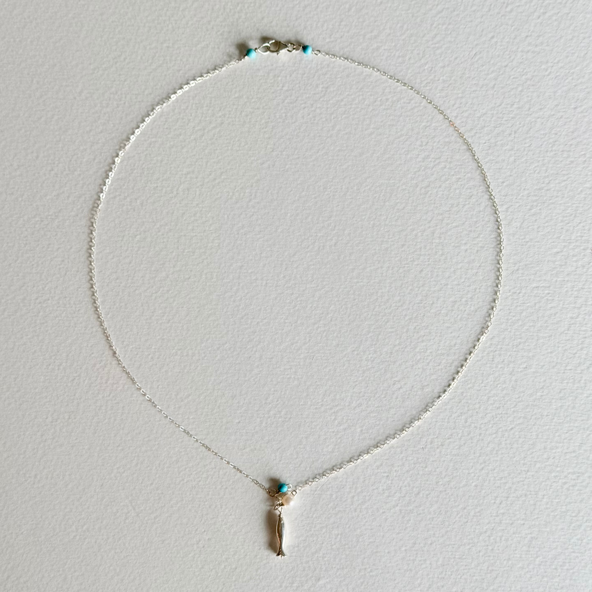 A delicate silver chain necklace with a small gold-toned pendant featuring a turquoise blue stone and a dangling gold charm, displayed on a textured white background.