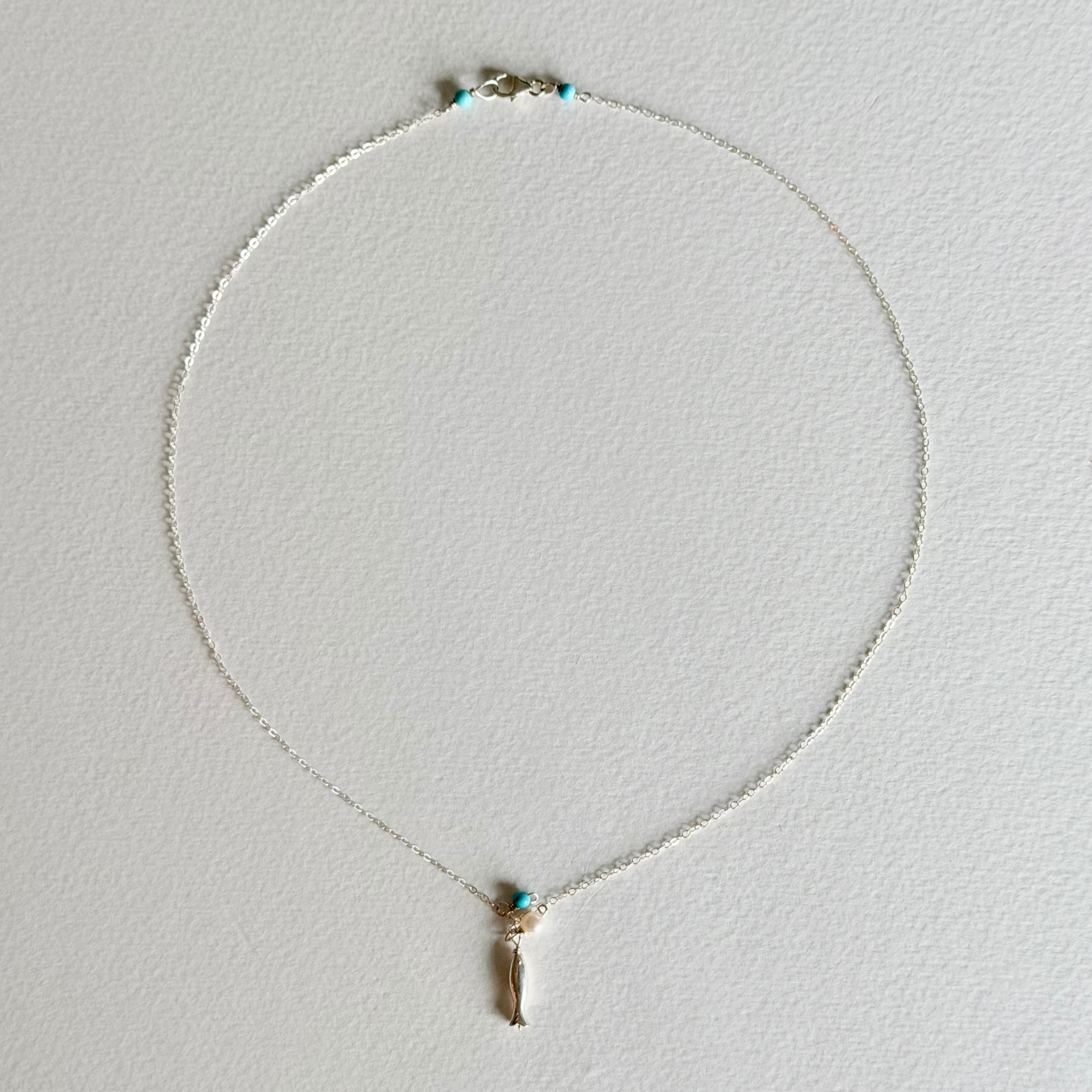 A delicate silver chain necklace with a small gold-toned pendant featuring a turquoise blue stone and a dangling gold charm, displayed on a textured white background.