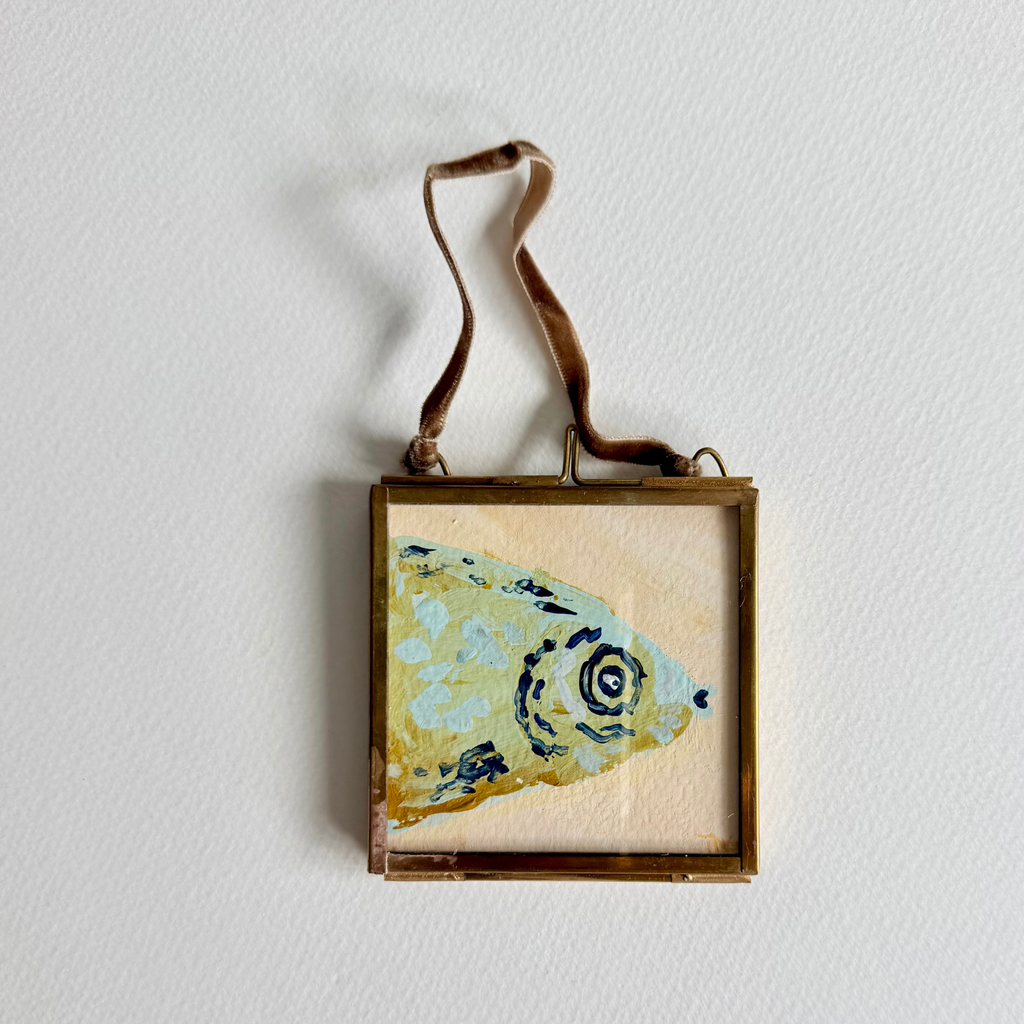 A square framed art piece featuring a textured painting of a fish head in light blue, gold, and dark blue, suspended by a brown ribbon against a white wall.