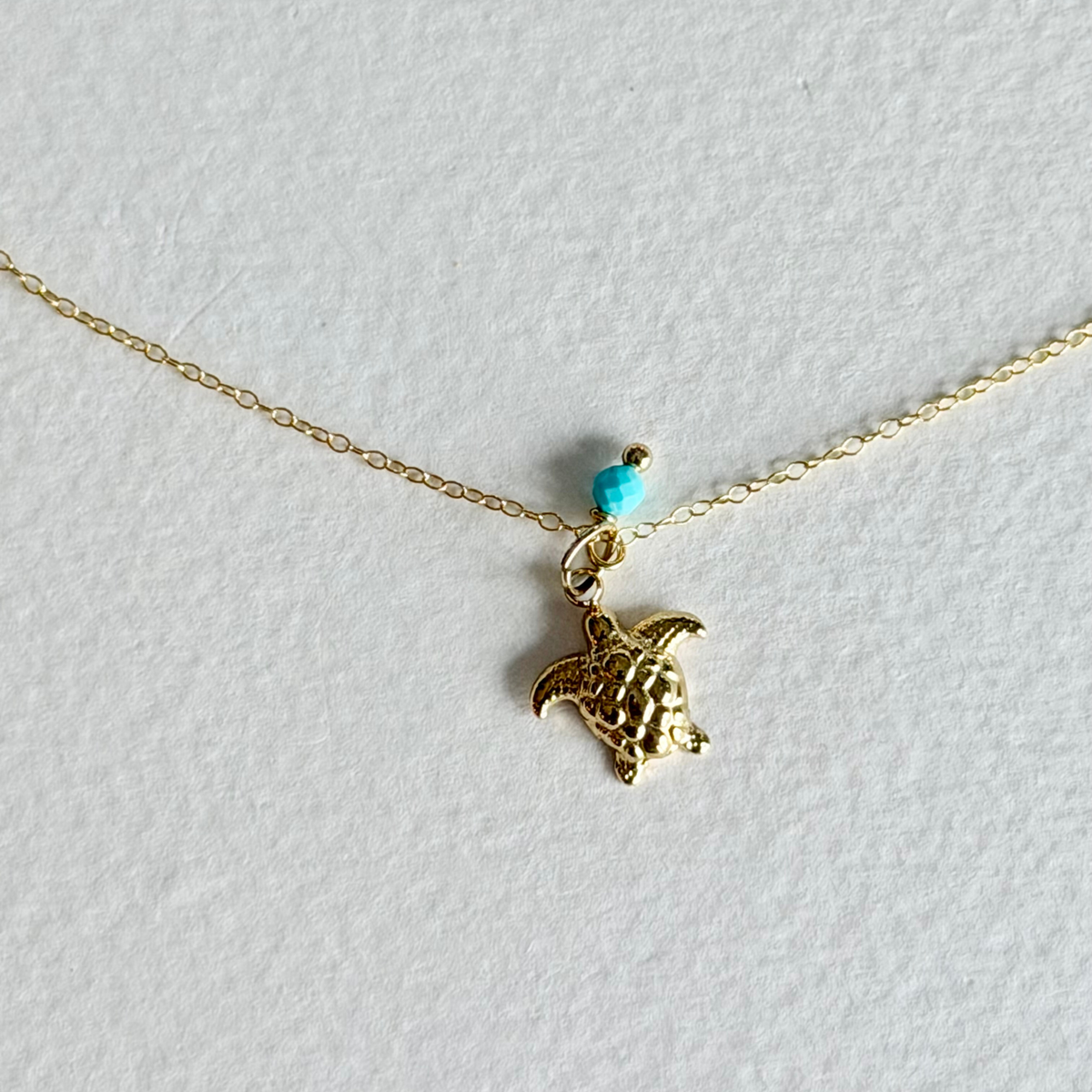 A delicate gold chain necklace featuring a small textured turtle pendant, a tiny turquoise bead, and a gold spacer, displayed on a textured white background.