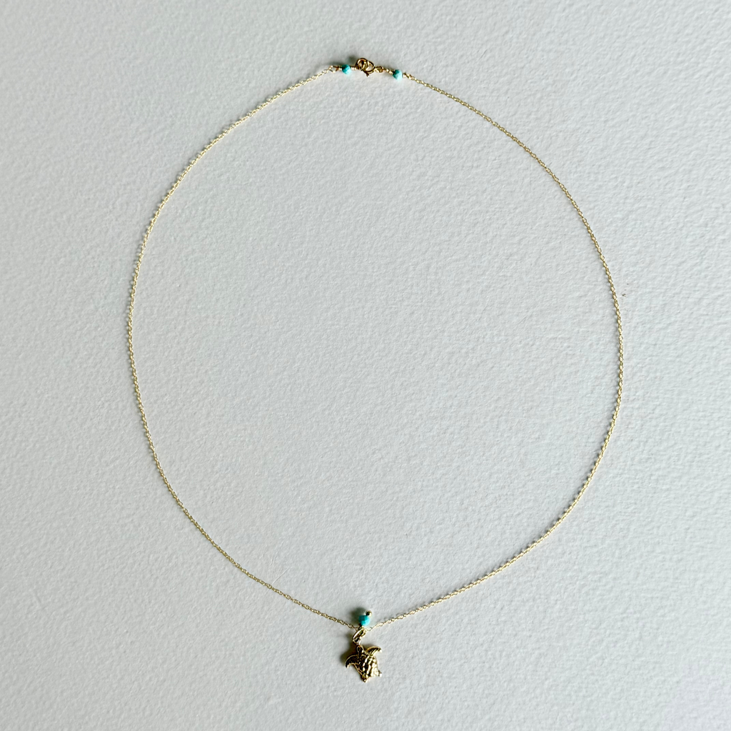A delicate gold chain necklace with a small gold bee pendant and tiny turquoise bead accents, displayed on a textured white background.