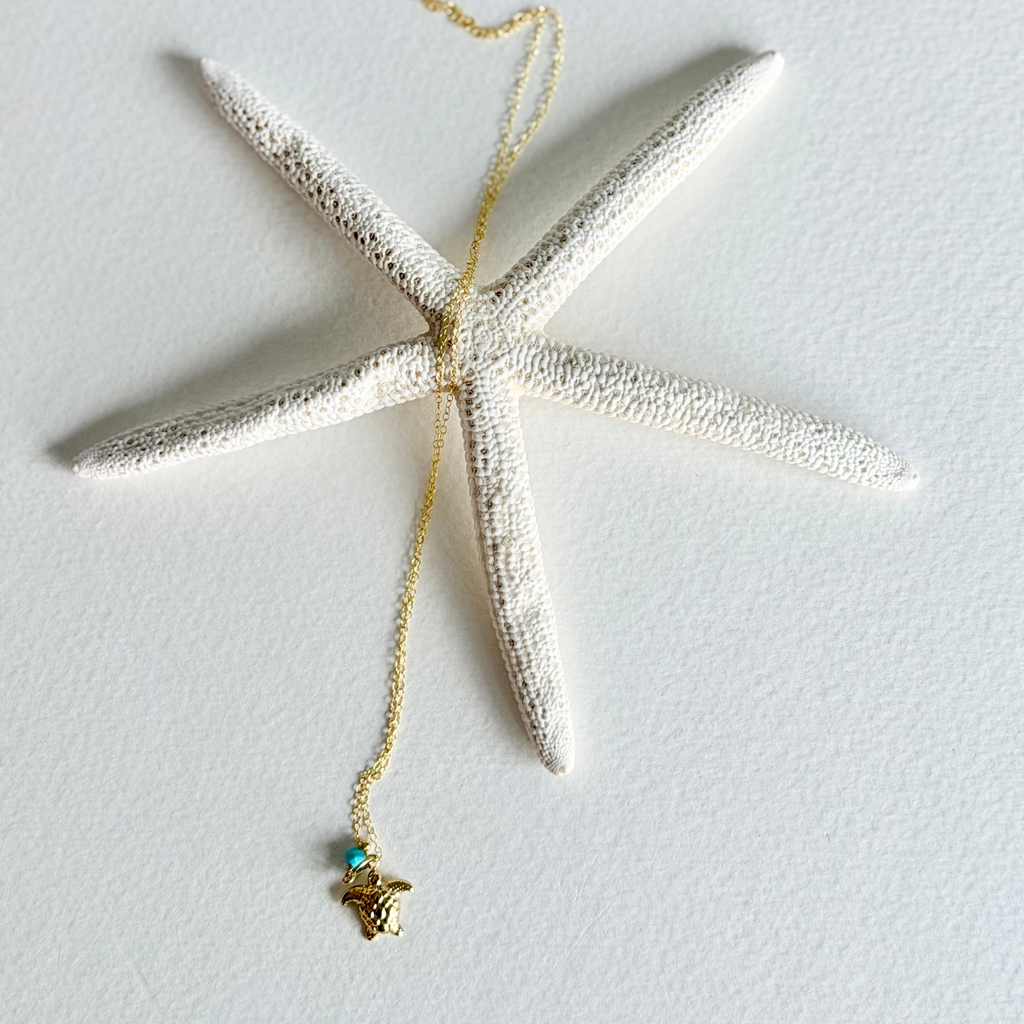 A gold necklace with a starfish-shaped pendant and a small turquoise bead, displayed on a textured white starfish prop against a white background.
