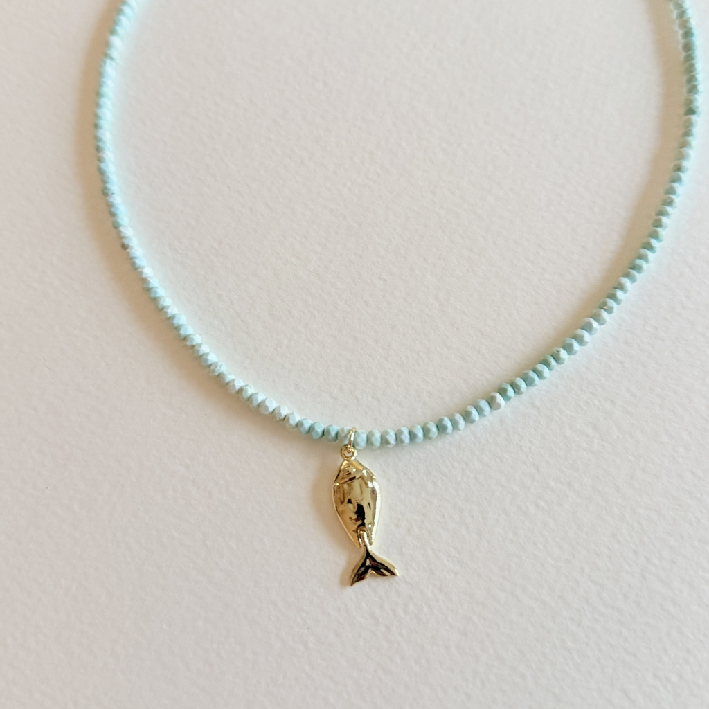 Close up view of Handmade aqua turquoise beaded necklace with a signature gold fish charm and 14k gold-filled clasp, measuring 16 inches with a 2-inch extender. Inspired by the colors of the sea and handcrafted by Marie & Oakley.