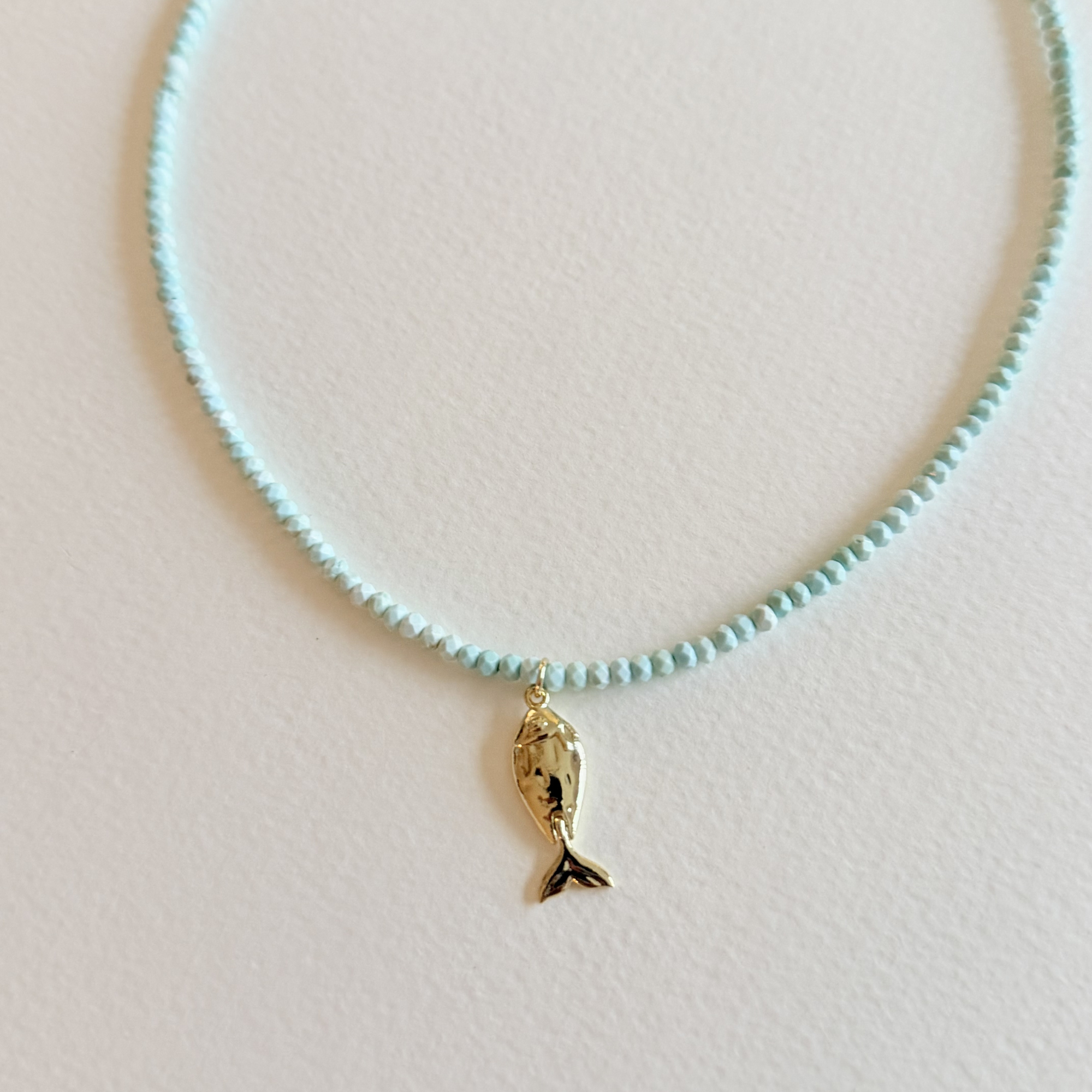 Close up view of Handmade aqua turquoise beaded necklace with a signature gold fish charm and 14k gold-filled clasp, measuring 16 inches with a 2-inch extender. Inspired by the colors of the sea and handcrafted by Marie & Oakley.