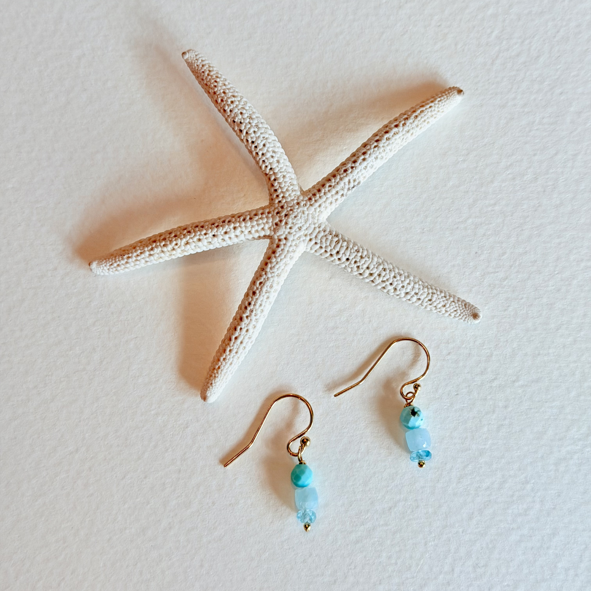 Pair of gold earrings with turquoise beads on a textured white surface with a starfish.