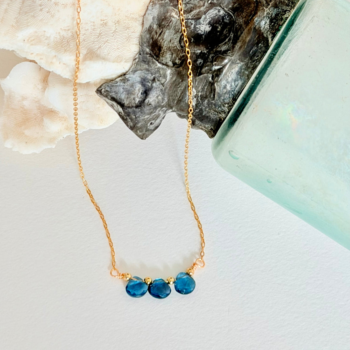 Gold necklace with blue beads on a white surface with a shell and crystal in the background