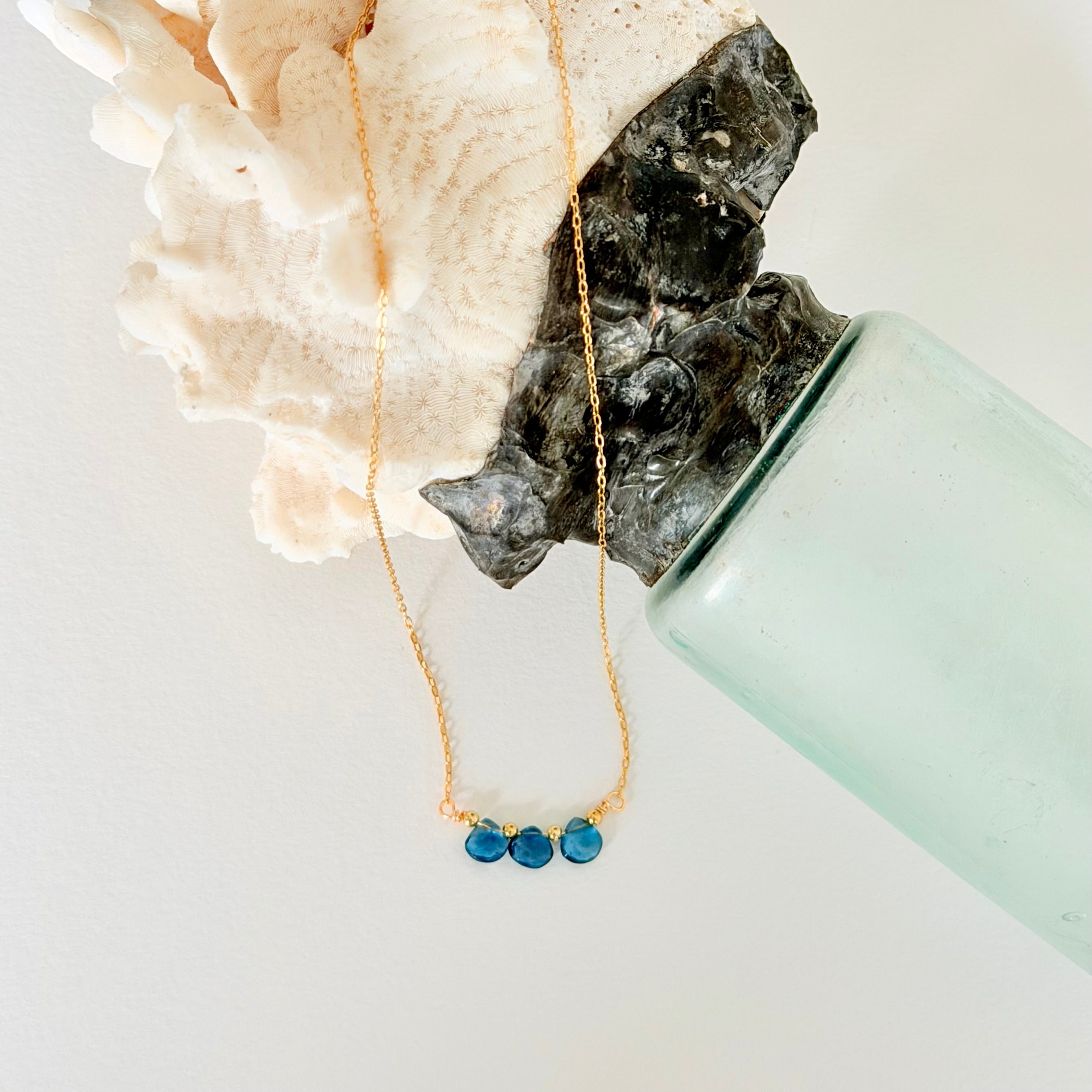 Gold necklace with blue beads on a white surface with a light green vase and white coral.