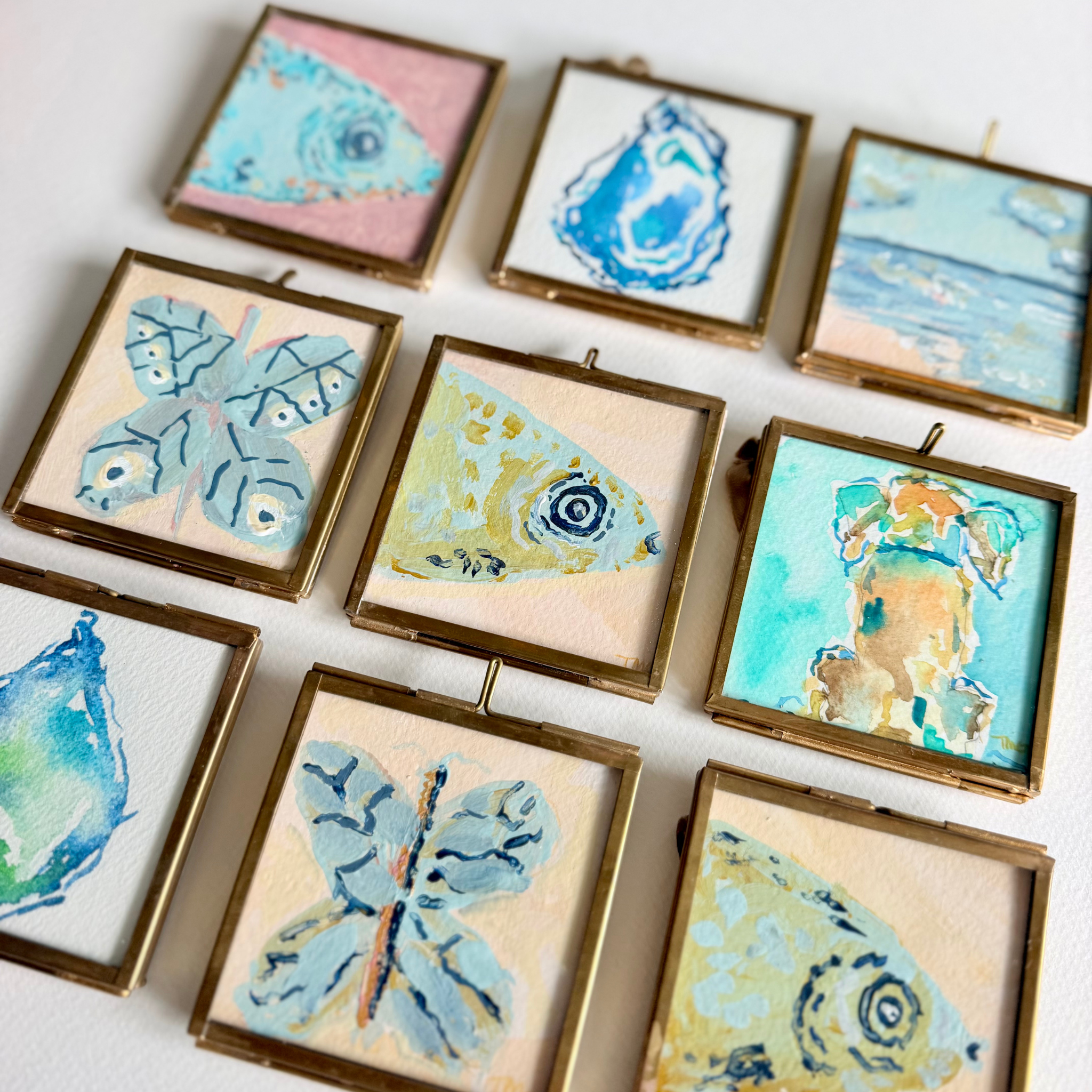 Nine small square framed artworks arranged on a white surface, each featuring a different painted nature motif such as a fish, butterfly, oyster, bird, and landscape. The frames appear metallic gold or brass, and the paintings use soft pastel and bright blue tones with cream, pink, and tan backgrounds.
