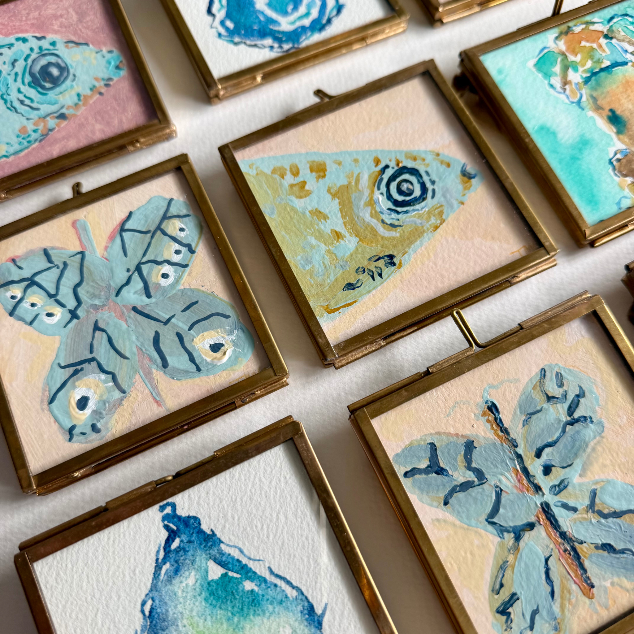 A close-up, angled view of several small square framed artworks arranged on a white surface. The pieces depict watercolor-style butterfly and fish illustrations in soft pastel tones including blue, teal, pink, and cream, each set in a gold-toned frame.