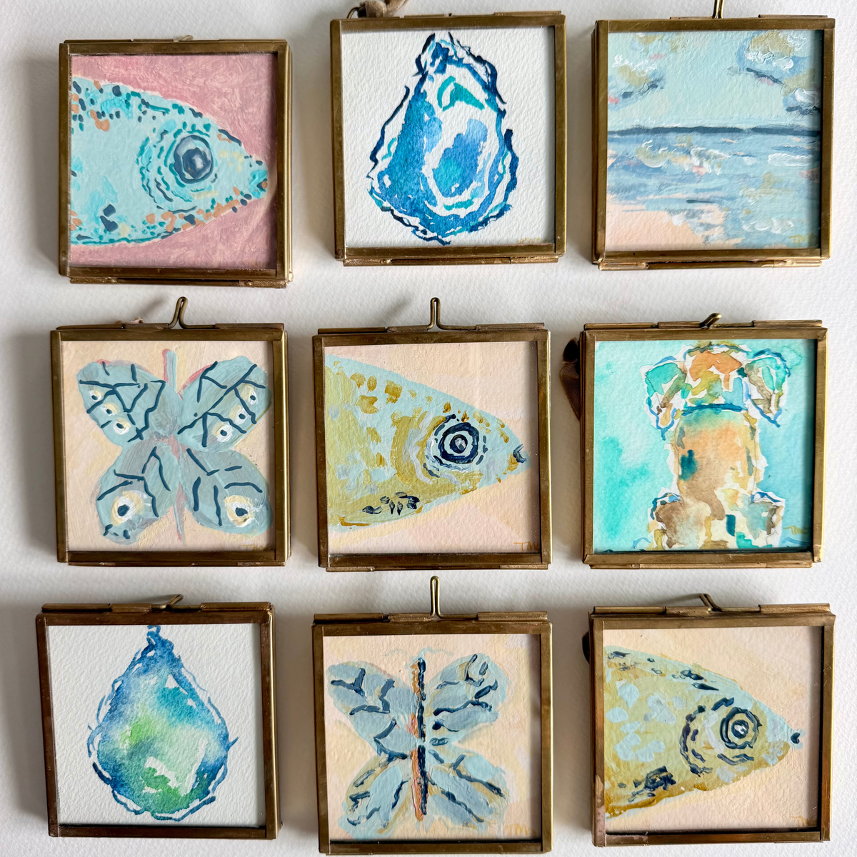 A 3x3 grid of small framed paintings on a white wall, each depicting a different nature-inspired design: fish, butterflies, a water droplet, and a beach scene. The paintings feature textured watercolor-style techniques in soft pastel tones of blue, teal, pink, beige, and yellow, housed in rustic gold-toned frames.