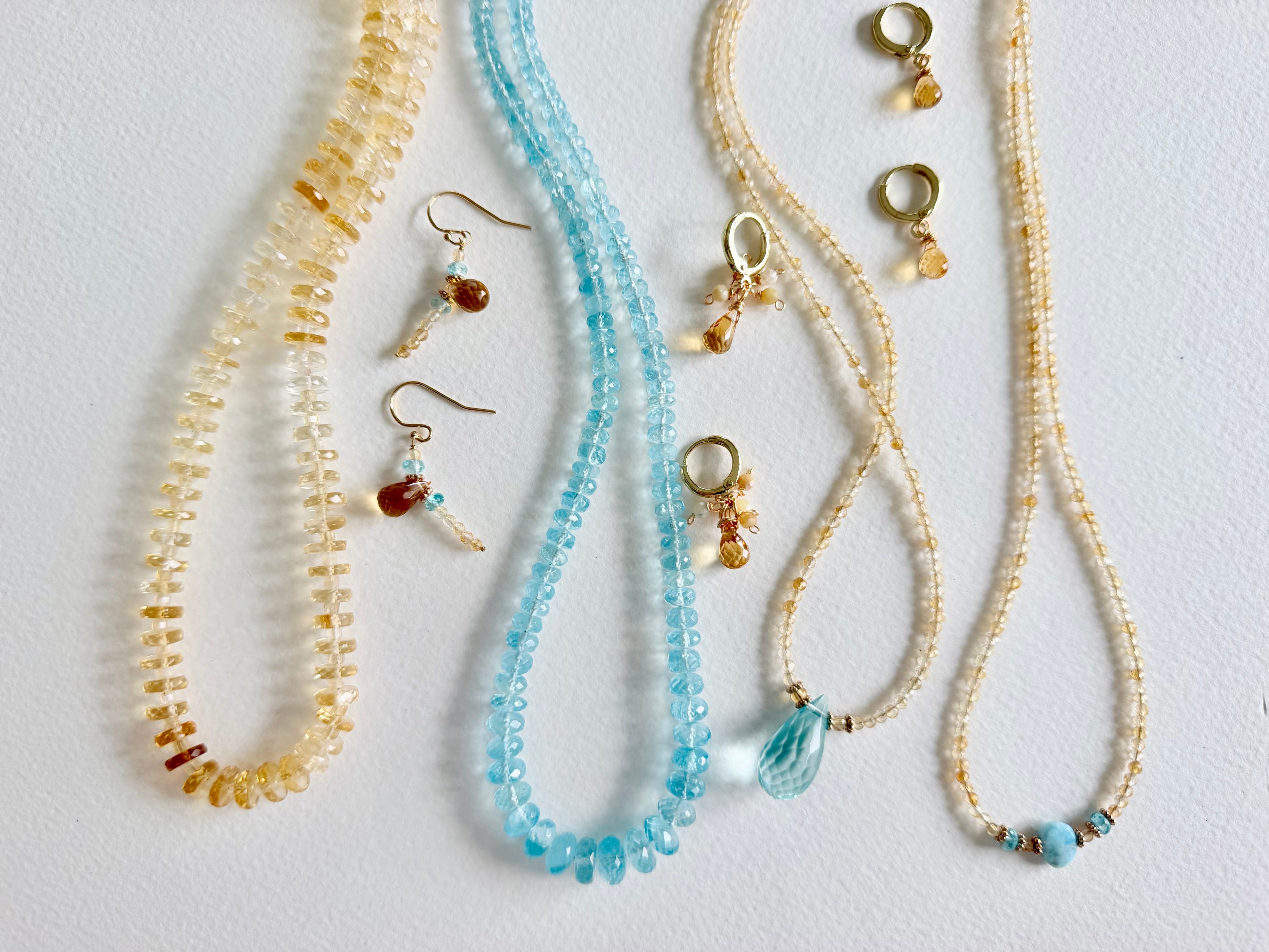 A set of handmade beaded jewelry laid out on a white surface, including two beaded necklaces, two beaded bracelets, and matching beaded earrings. The necklaces and earrings feature light blue beads, while the bracelets feature pale gold beads.