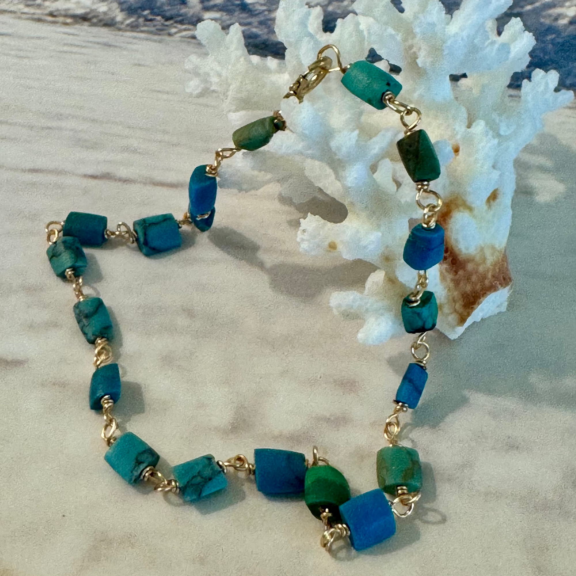 A bracelet made of genuine matte turquoise beads with a smooth, flat varying in intensity of green to blue on 14k gold filled wire. Displayed against white coral on a beachy background.