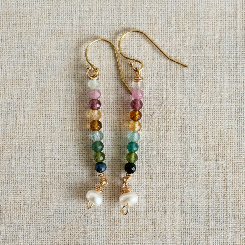 Multicolor beaded dangle earrings with pearl at the end on 14k gold filled wire displayed against a textured sand white background.