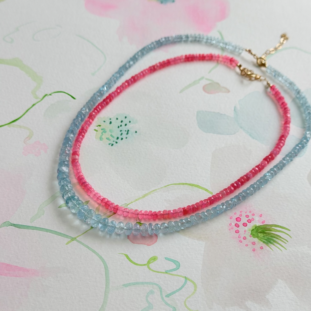 Two beaded necklaces, one pink and one blue, on a floral-patterned fabric background.
