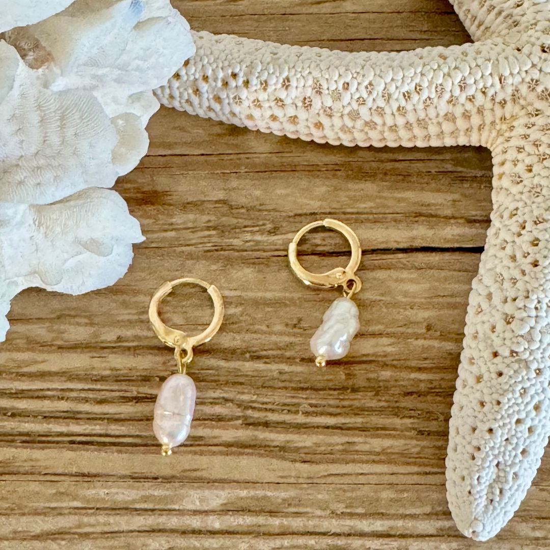 Pair of earrings with a single oblong freshwater pearl on each hoop handcrafted with 14k gold-filled wire huggie clasp. Displayed between creamy white coral and starfish on a beachy wood grain surface.