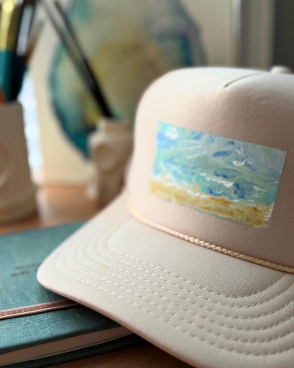Cream hand-painted trucker hat featuring original textured ocean artwork in turquoise, blue and sandy gold by Marie & Oakley.