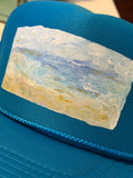 A close-up of a bright blue trucker hat featuring an impressionistic painting of a beach scene in shades of blue, white, and beige.