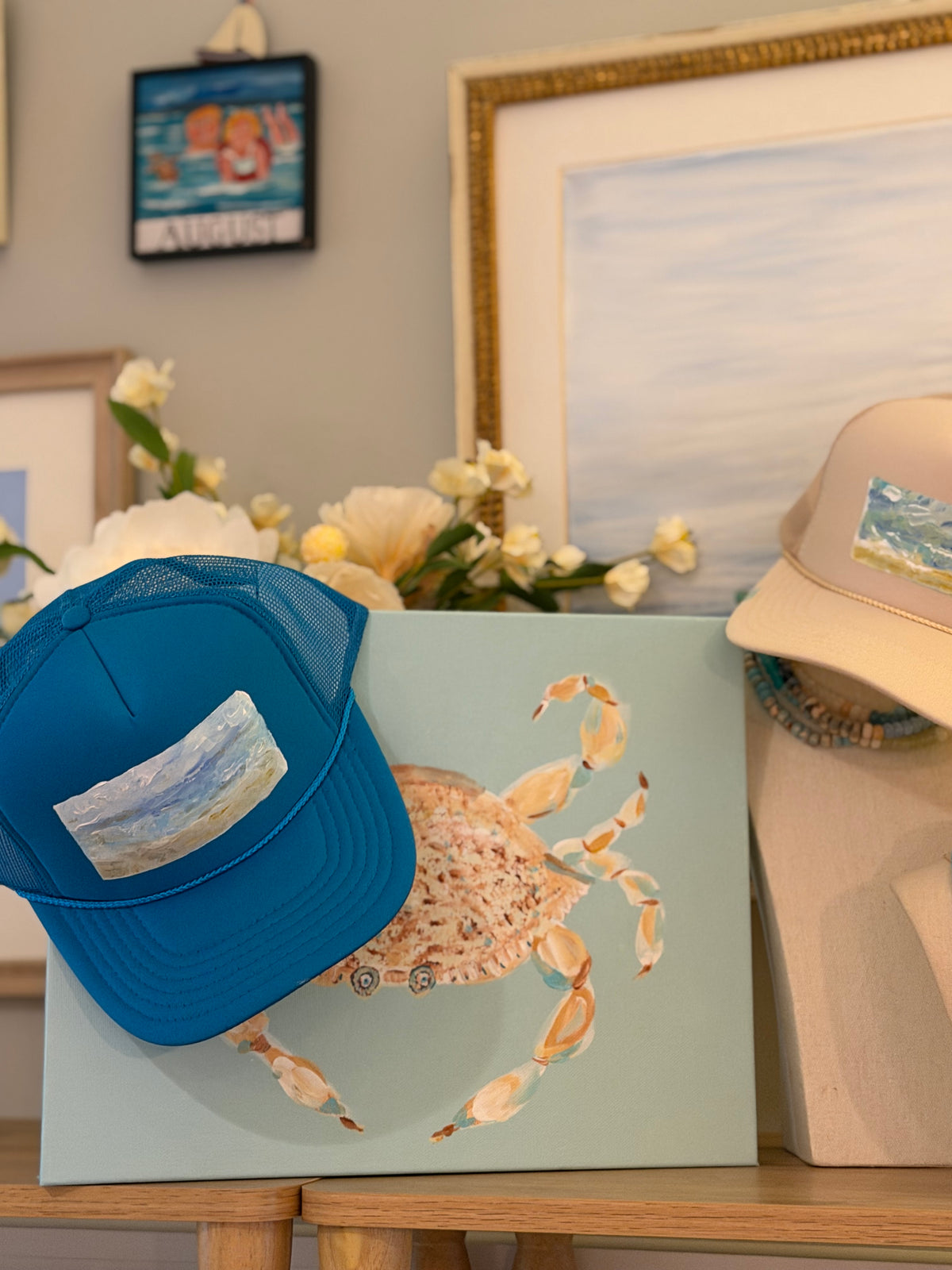 A blue trucker hat with a rectangular beach scene patch is draped over a canvas painting of a crab on a light blue background, styled on a wooden table with a beige hat and framed art in the background.