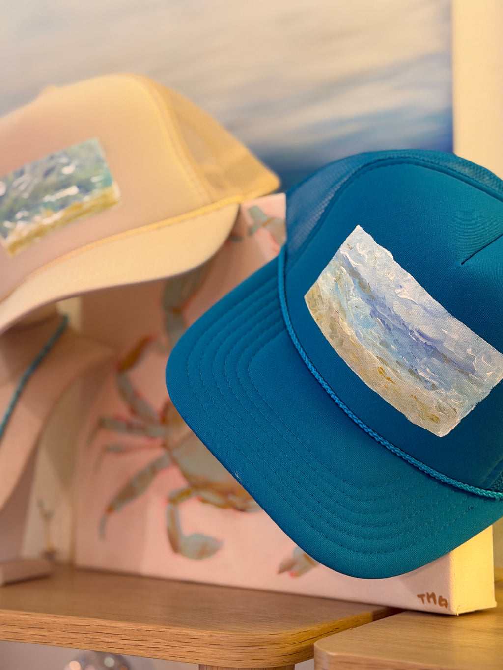 A teal blue trucker hat with a rectangular patch depicting a beach scene, displayed on a wooden shelf next to a cream-colored hat.