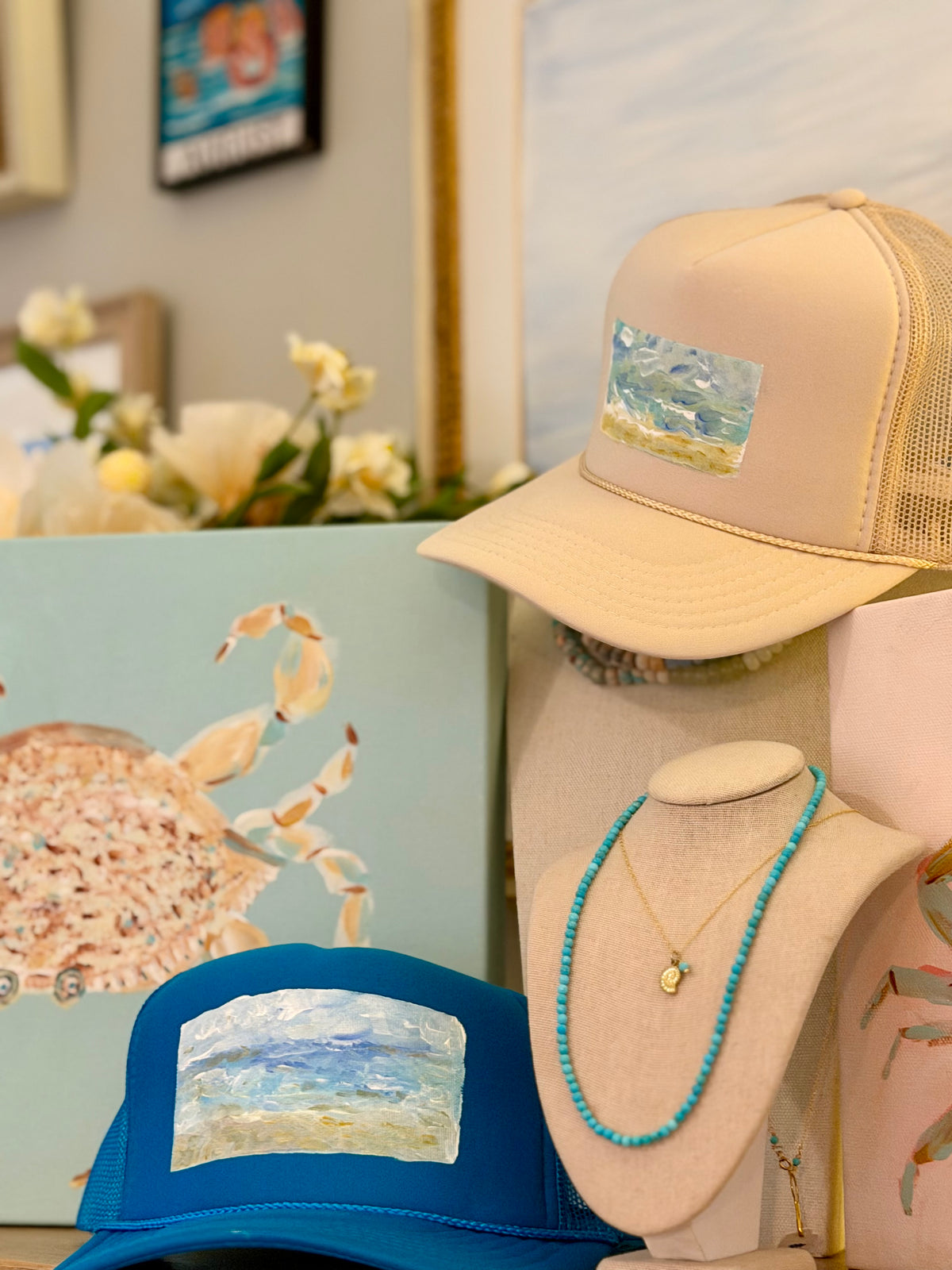 A styled display of beach-themed merchandise, featuring two trucker hats with matching canvas prints of a beach scene, a turquoise beaded necklace on a cream bust, and a light blue canvas print with a crab illustration.