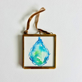 A square framed watercolor art painting featuring a blue and green teardrop design on a white background, enclosed in a gold-toned frame with a brown hanging ribbon, shown against a white wall.