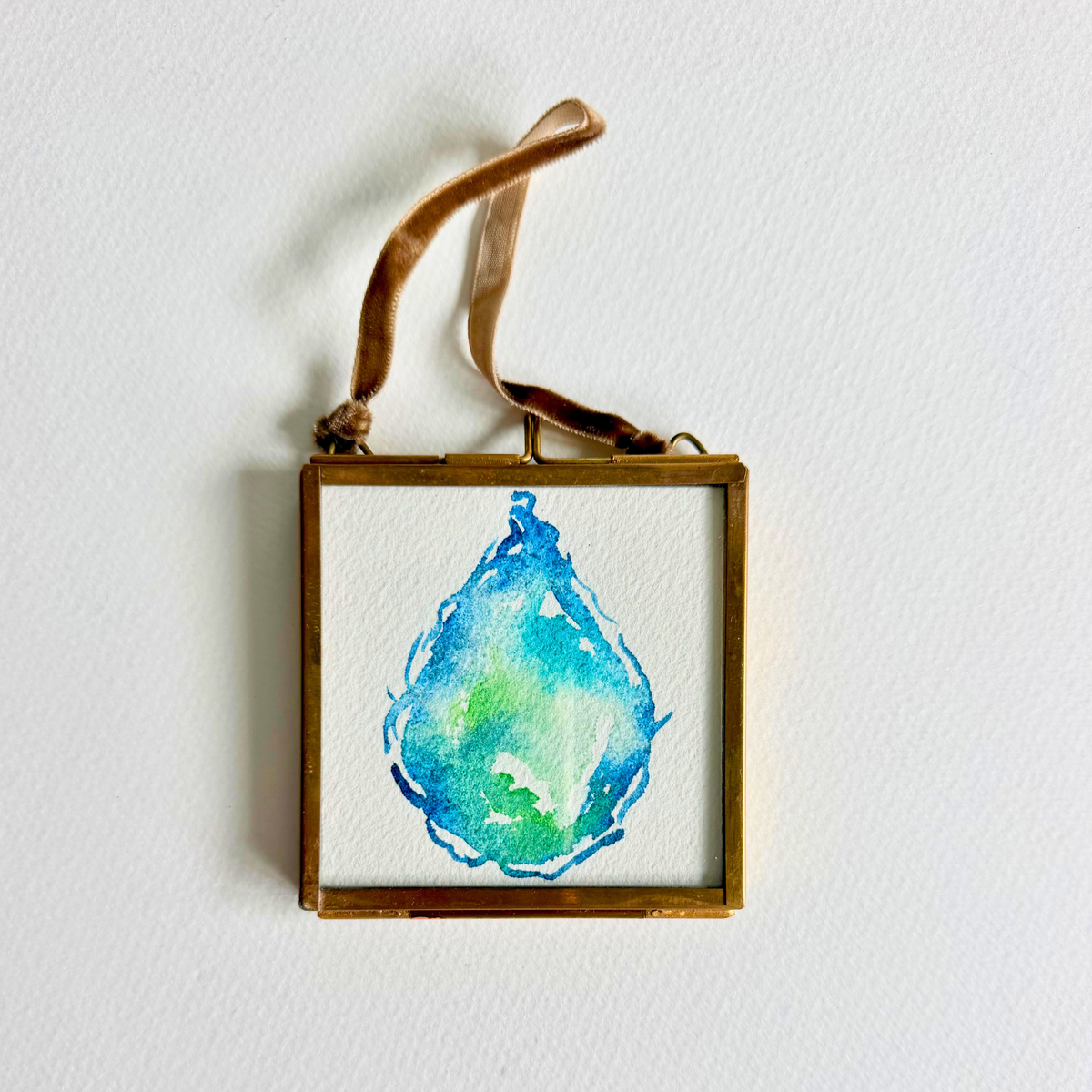 A square framed watercolor art painting featuring a blue and green teardrop design on a white background, enclosed in a gold-toned frame with a brown hanging ribbon, shown against a white wall.