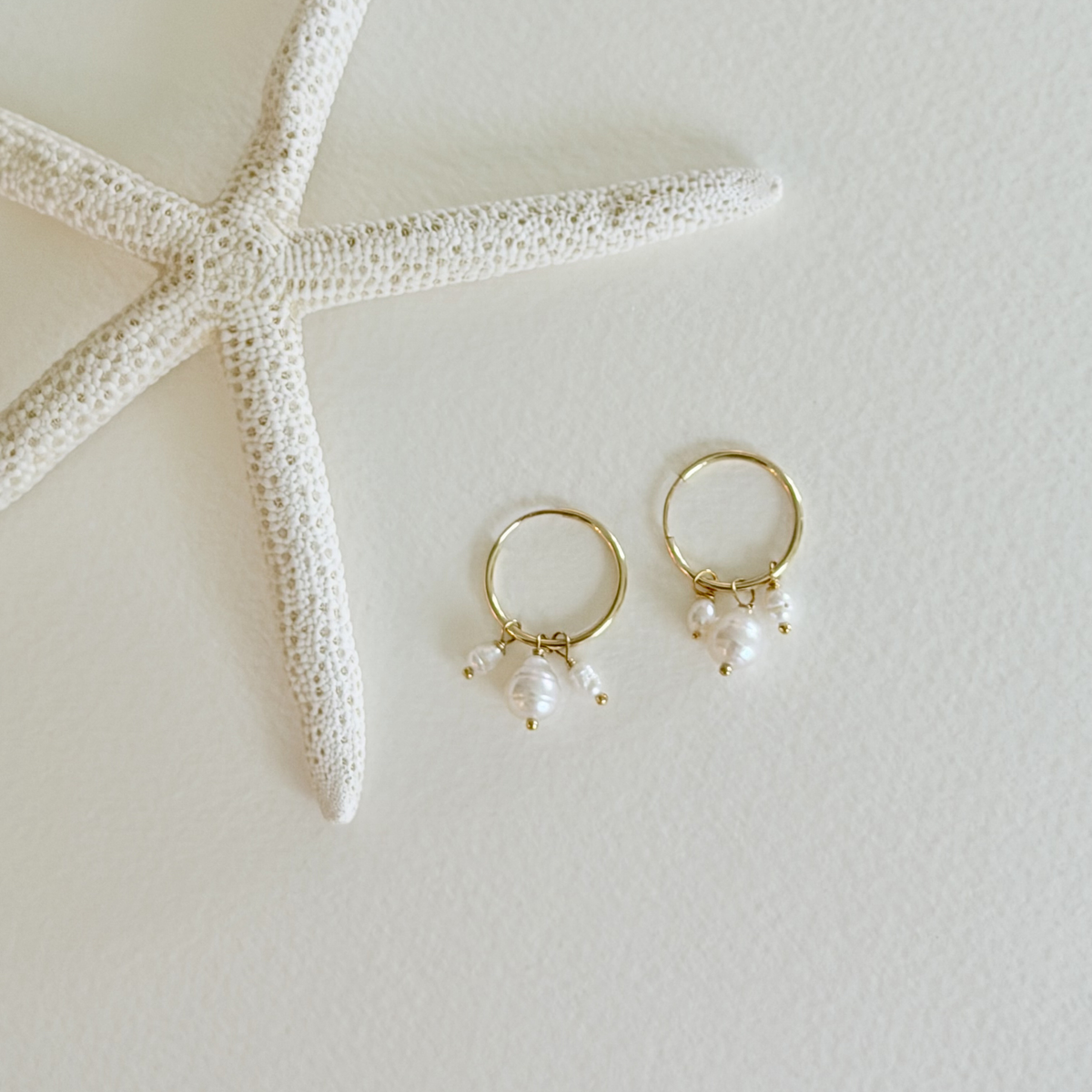 Gold hoop earrings with pearls on a light surface next to a starfish.