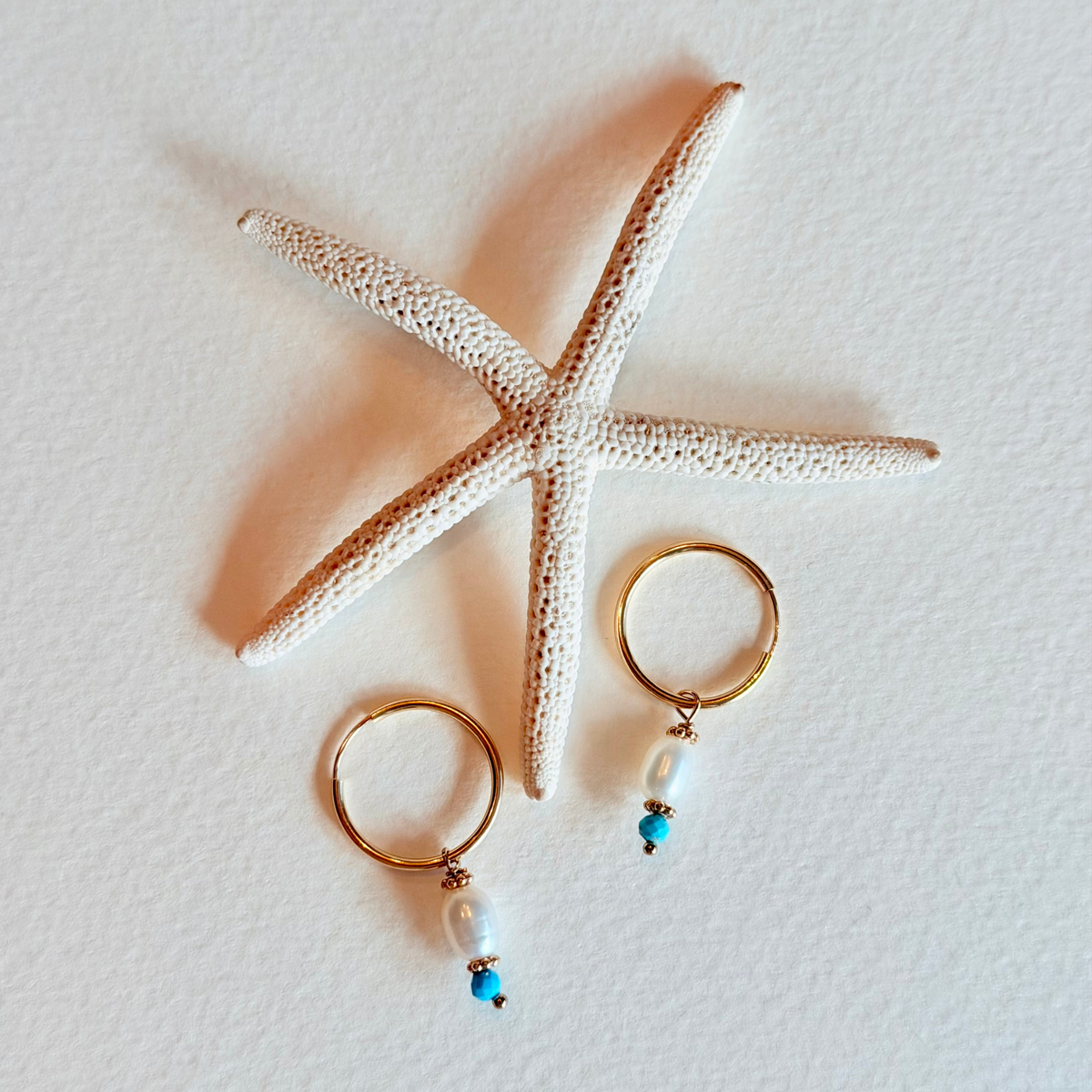 Gold hoop earrings with pearls and turquoise beads on a starfish and white textured background