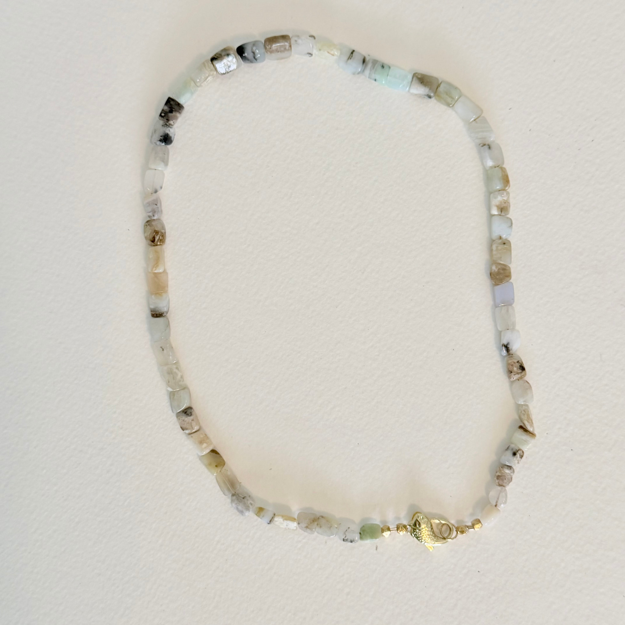 A beaded necklace made of small, irregular natural stones in shades of white, gray, and light green, featuring a small gold clasp, displayed on a textured off-white background.