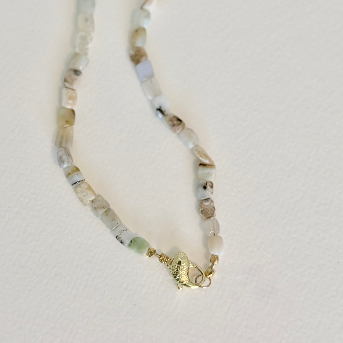 A beaded necklace featuring small, irregular natural stone beads in muted shades of beige, gray, and soft green, accented by a small gold fish charm and gold clasp, displayed on a textured off-white background.