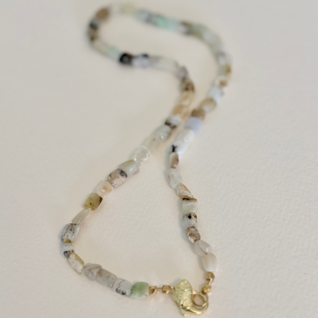 A beaded necklace made of small natural stones in white, beige, light green, and gray, with a gold clasp, photographed on a plain off-white background.