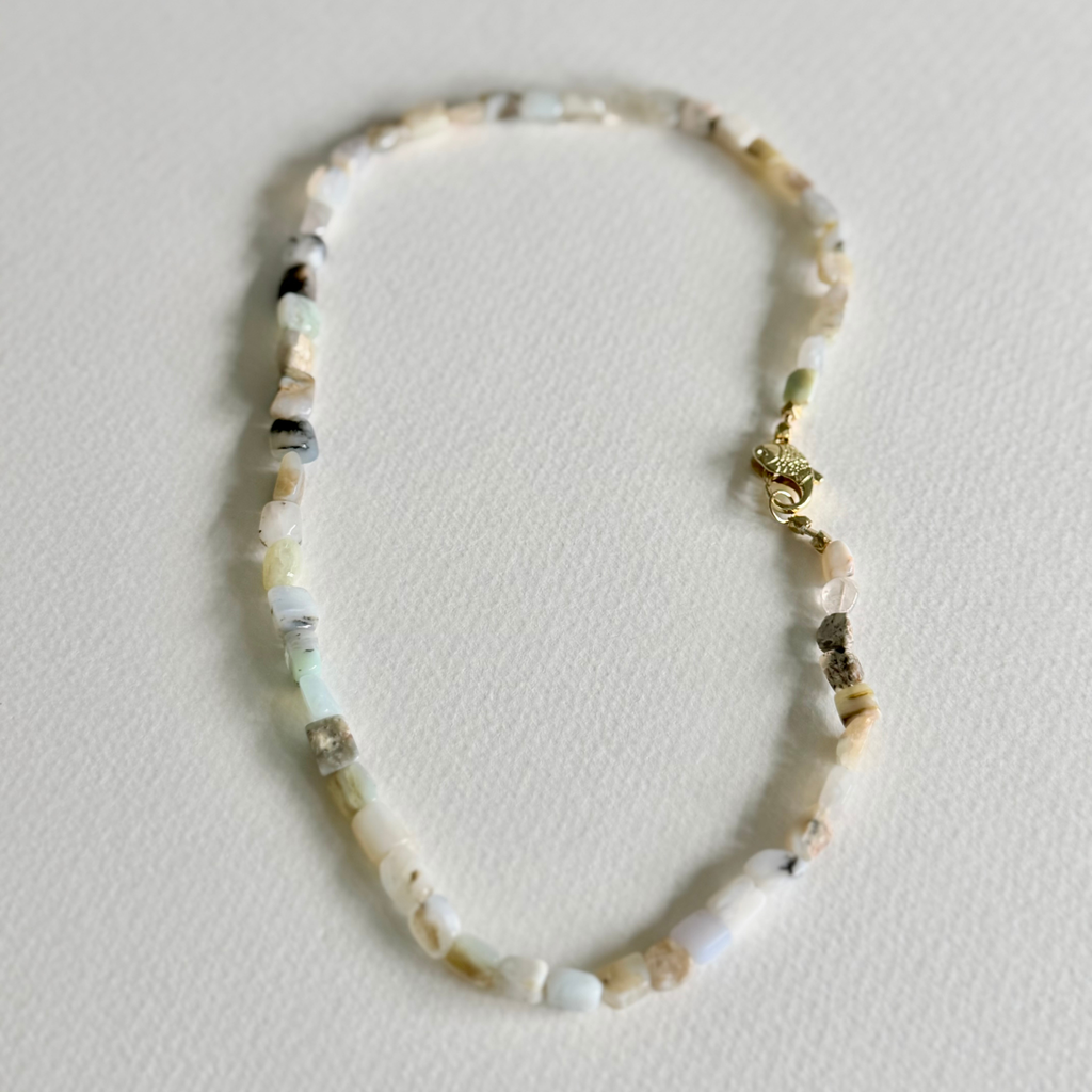 A delicate beaded necklace made of small, irregular natural stone chips in off-white, cream, and light gray, featuring a small gold lobster clasp, displayed on a textured off-white surface.