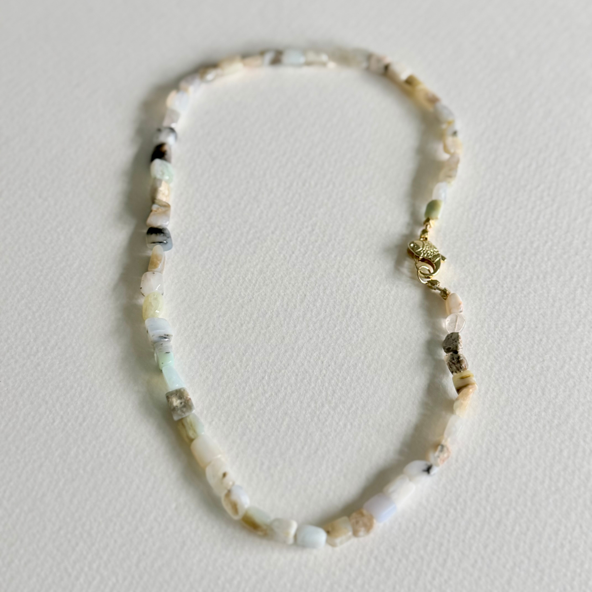 A delicate beaded necklace made of small, irregular natural stone chips in off-white, cream, and light gray, featuring a small gold lobster clasp, displayed on a textured off-white surface.