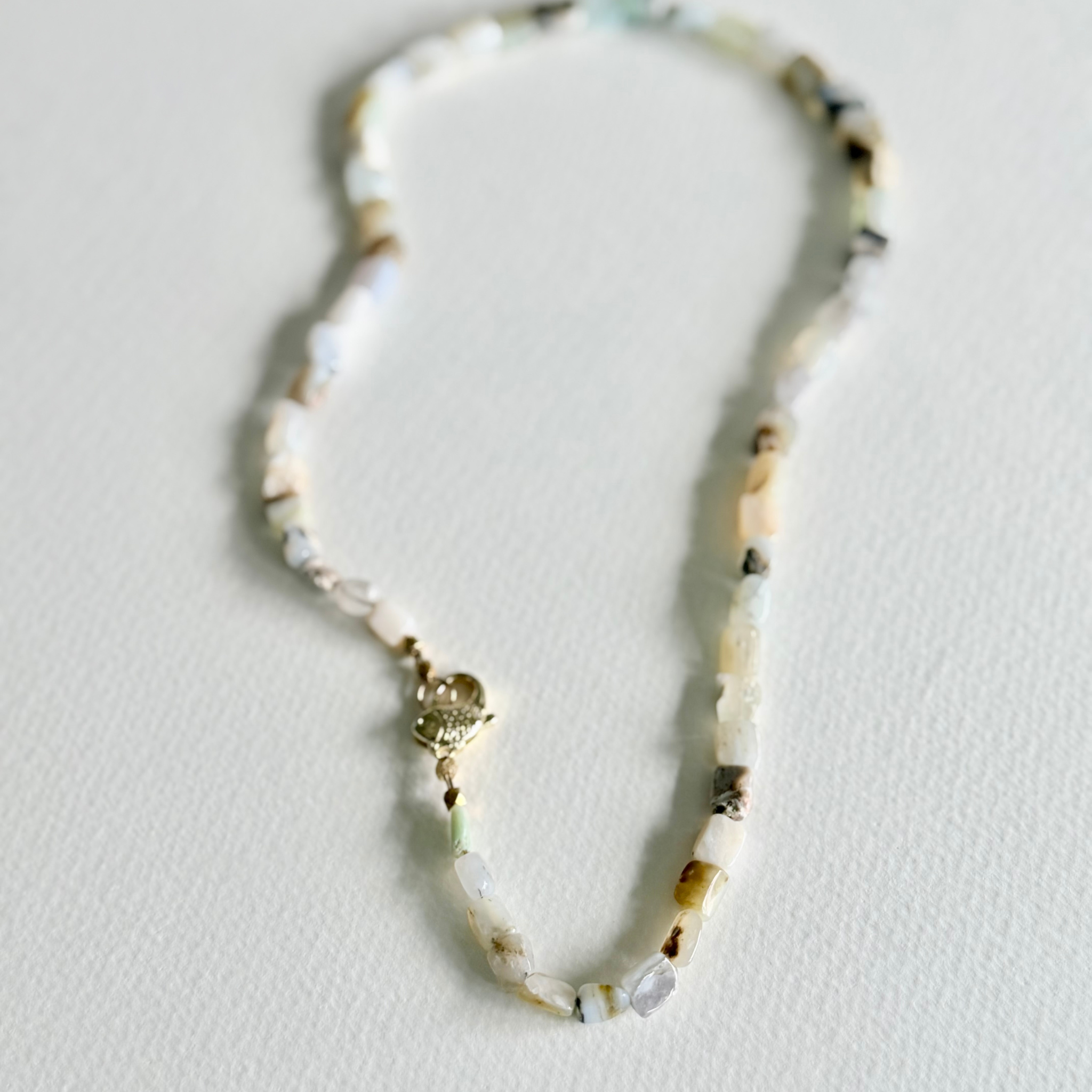 A beaded necklace made of small irregular shell-like pieces in off-white, cream, tan, and gray, with a small gold clasp, photographed on a plain light background.