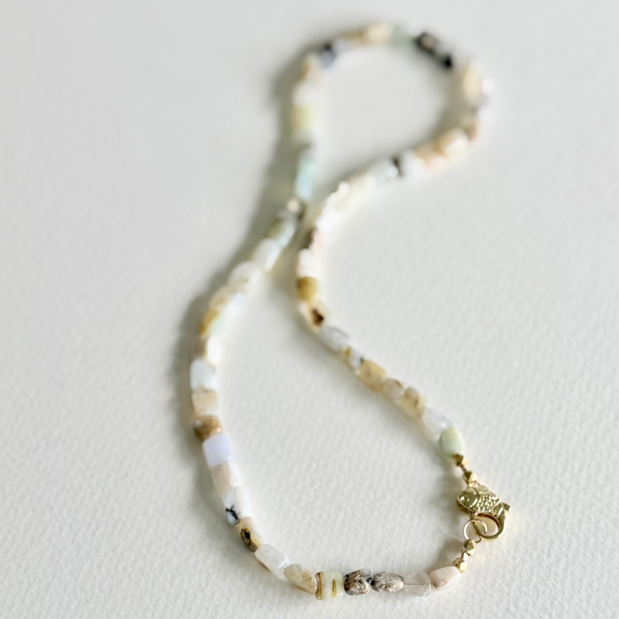 A beaded necklace made of small irregular stone beads in cream, beige, white, and pale green tones, with a gold-toned clasp, photographed on a plain light background.