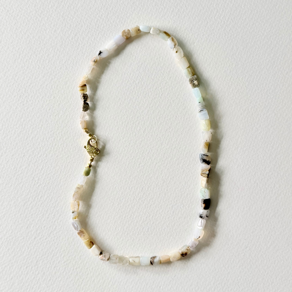 A beaded necklace made of small natural stone beads in cream, white, light green, and brown, with a small gold clasp, laid flat on a textured off-white background.