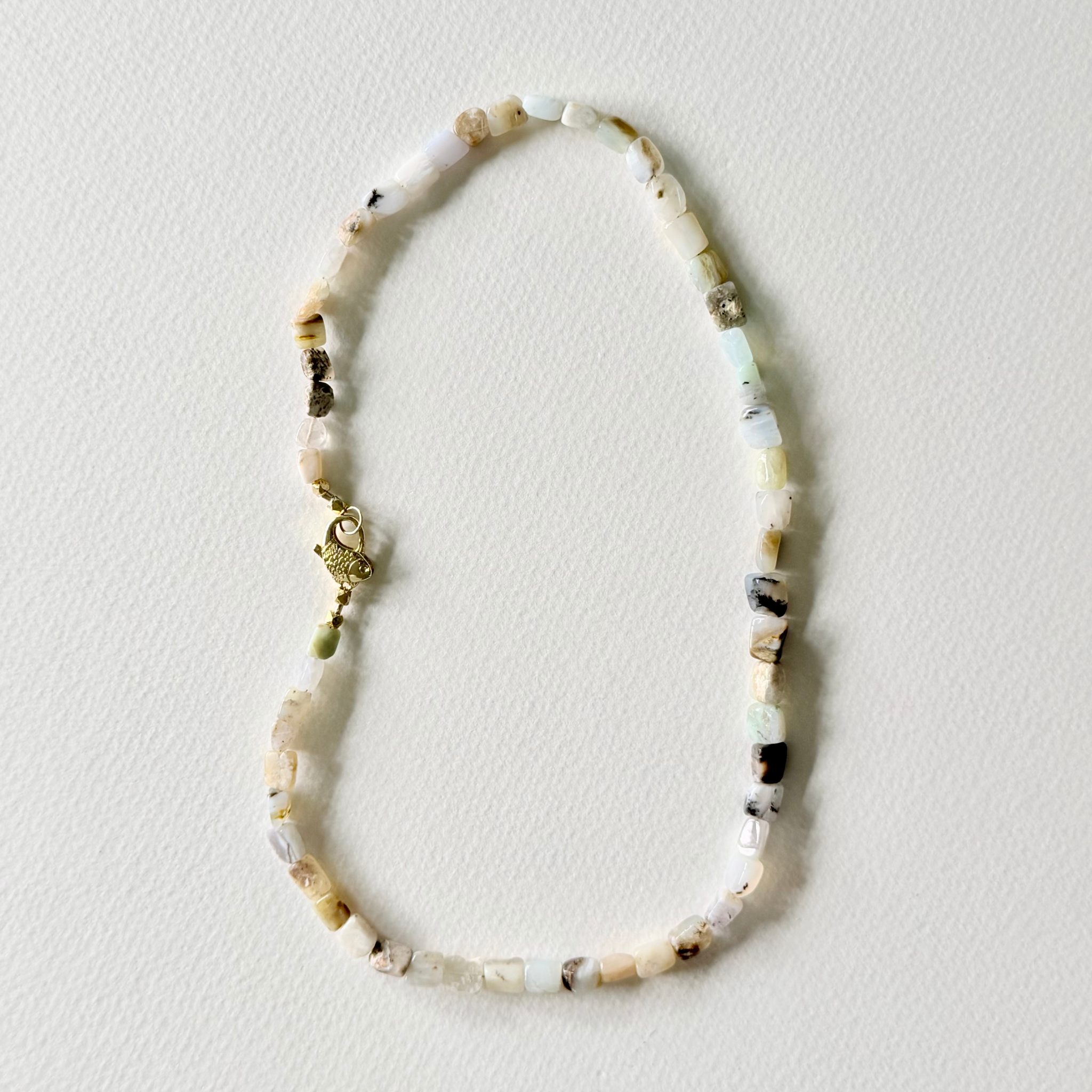A beaded necklace made of small natural stone beads in cream, white, light green, and brown, with a small gold clasp, laid flat on a textured off-white background.