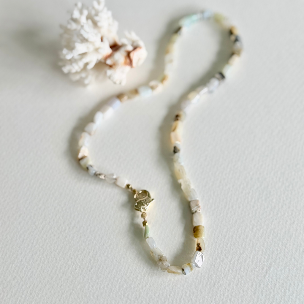A beaded necklace made of small natural Peruvian Opal beads in soft cream, beige, white, and muted green tones, with a small gold clasp, photographed on a light neutral surface with a decorative coral-like prop.