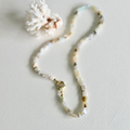 A beaded necklace made of small natural Peruvian Opal beads in soft cream, beige, white, and muted green tones, with a small gold clasp, photographed on a light neutral surface with a decorative coral-like prop.