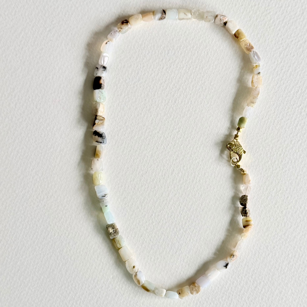 A beaded necklace made of irregular, chunky stone beads in cream, white, and light brown, featuring a small gold-toned elephant charm and clasp, laid flat on a textured off-white background.