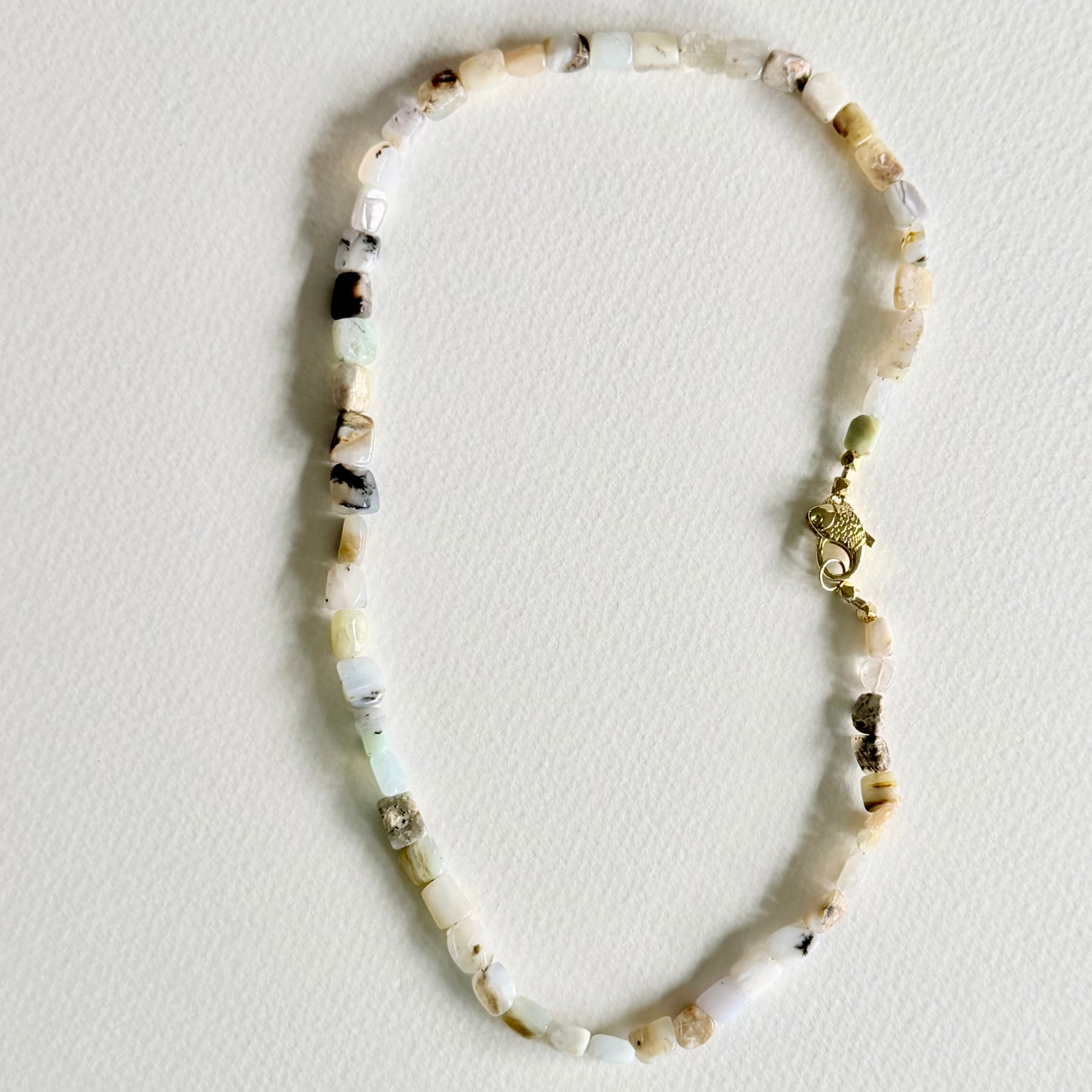 A beaded necklace made of irregular, chunky stone beads in cream, white, and light brown, featuring a small gold-toned elephant charm and clasp, laid flat on a textured off-white background.